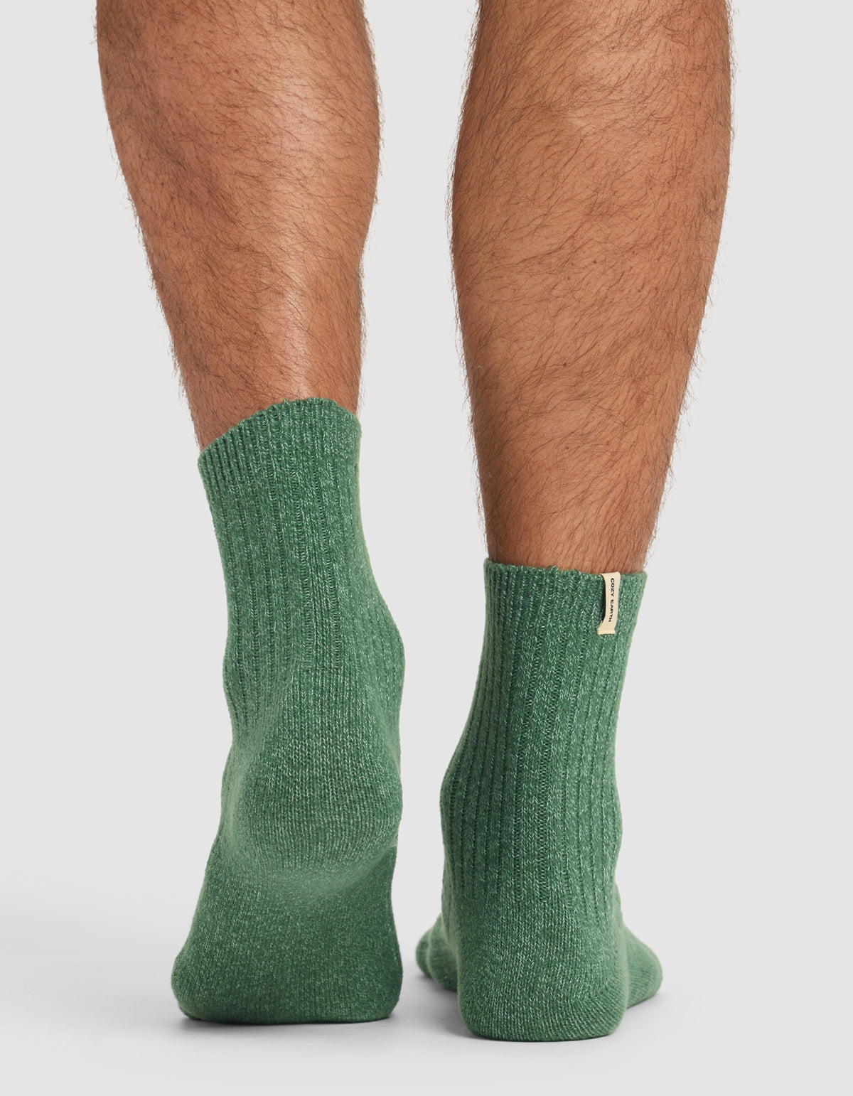 A close-up of someone wearing Cozy Earth’s The Plush Modern Crew Sock 3-Pack in green, showing ribbed texture and a small fabric tag on the cuff, standing on a flat surface.