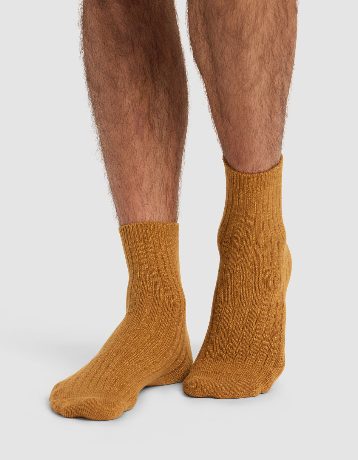 Someone with hairy legs is wearing The Plush Modern Crew Sock 3-Pack by Cozy Earth in mustard yellow, featuring a ribbed knit design, standing against a plain white background.