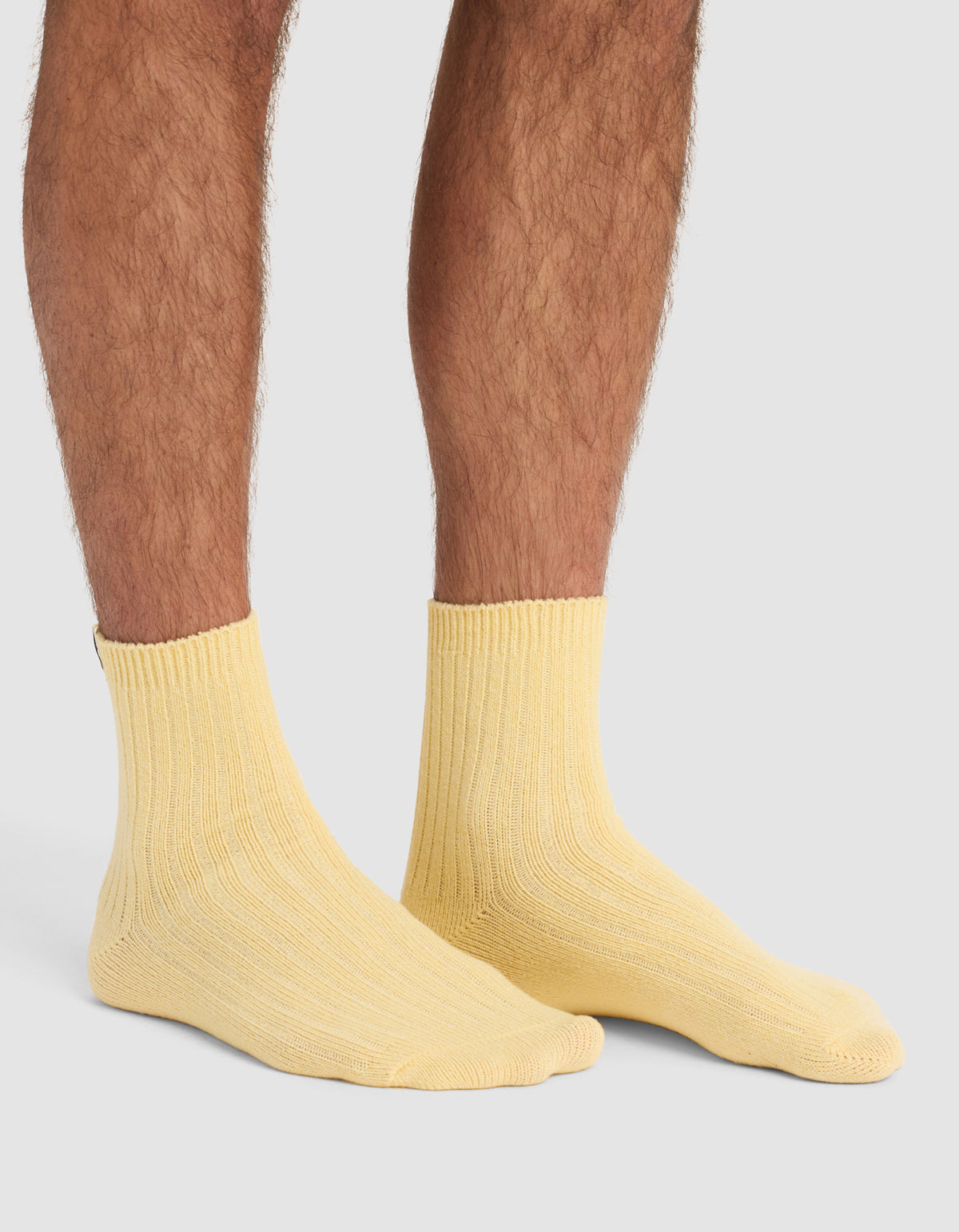 A person wears The Plush Modern Crew Sock 3-Pack by Cozy Earth in light yellow, standing on a plain light gray background. Only their lower legs and feet are visible.