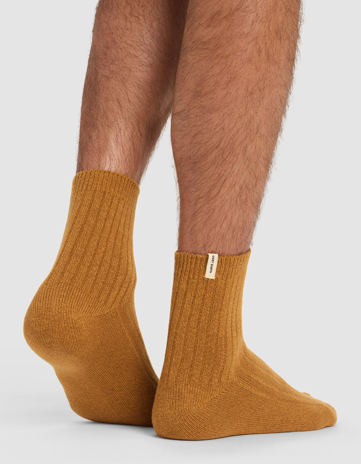 A close-up of someone wearing Cozy Earth’s The Plush Modern Crew Sock 3-Pack in ribbed mustard, featuring a small fabric tag, standing on a light grey surface.