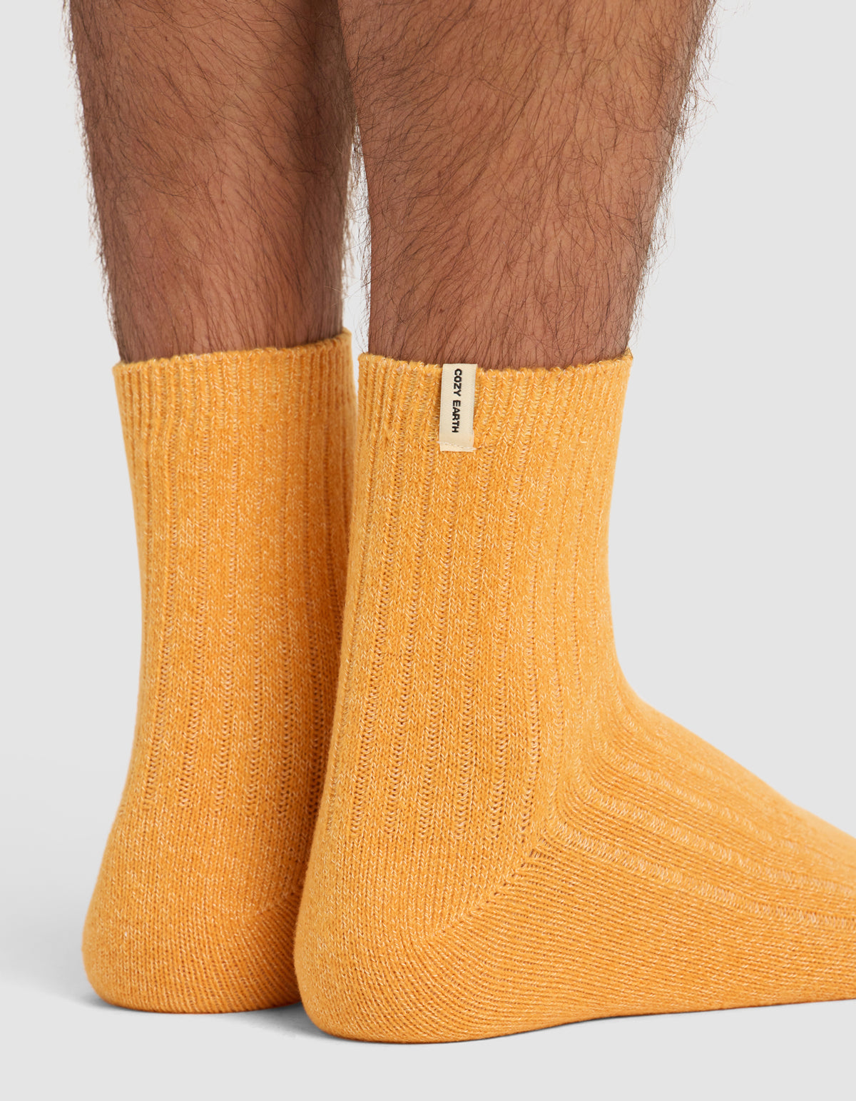 Close-up of lower legs wearing The Plush Modern Crew Sock by Cozy Earth in mustard-yellow, featuring ribbed design and a beige label on the cuff, shown on a light gray background.