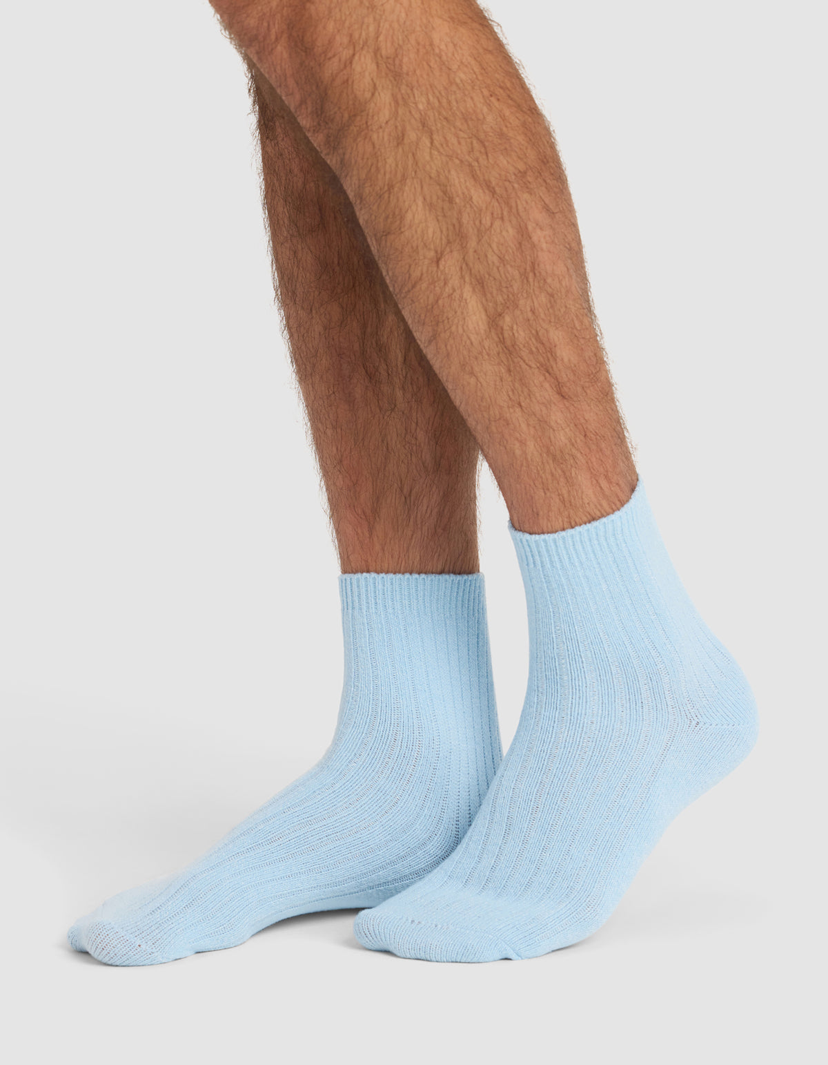 A person with crossed legs is wearing Cozy Earth’s The Plush Modern Crew Sock 3-Pack in light blue, featuring a ribbed crew design against a plain white background.