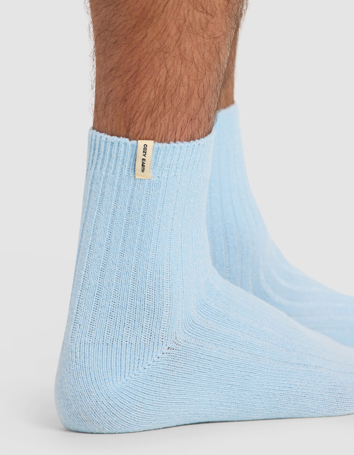 A close-up side view of a person wearing The Plush Modern Crew Sock by Cozy Earth in light blue, featuring a ribbed texture and small beige tag, against a plain white background.