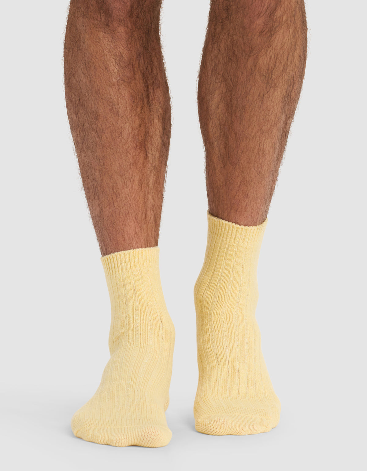 A person stands on a light surface, shown from the calves down, wearing Cozy Earth's The Plush Modern Crew Sock in pale yellow. Their feet are close together, set against a neutral gray background.