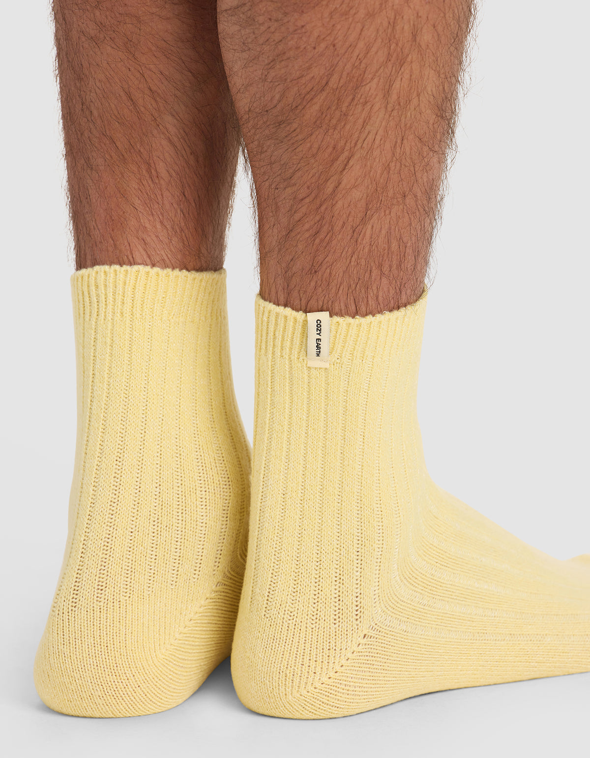 Close-up of a person wearing Cozy Earth’s The Plush Modern Crew Sock in light yellow, standing on a light surface. A small white tag is visible near the ankle, and leg hair can be seen.