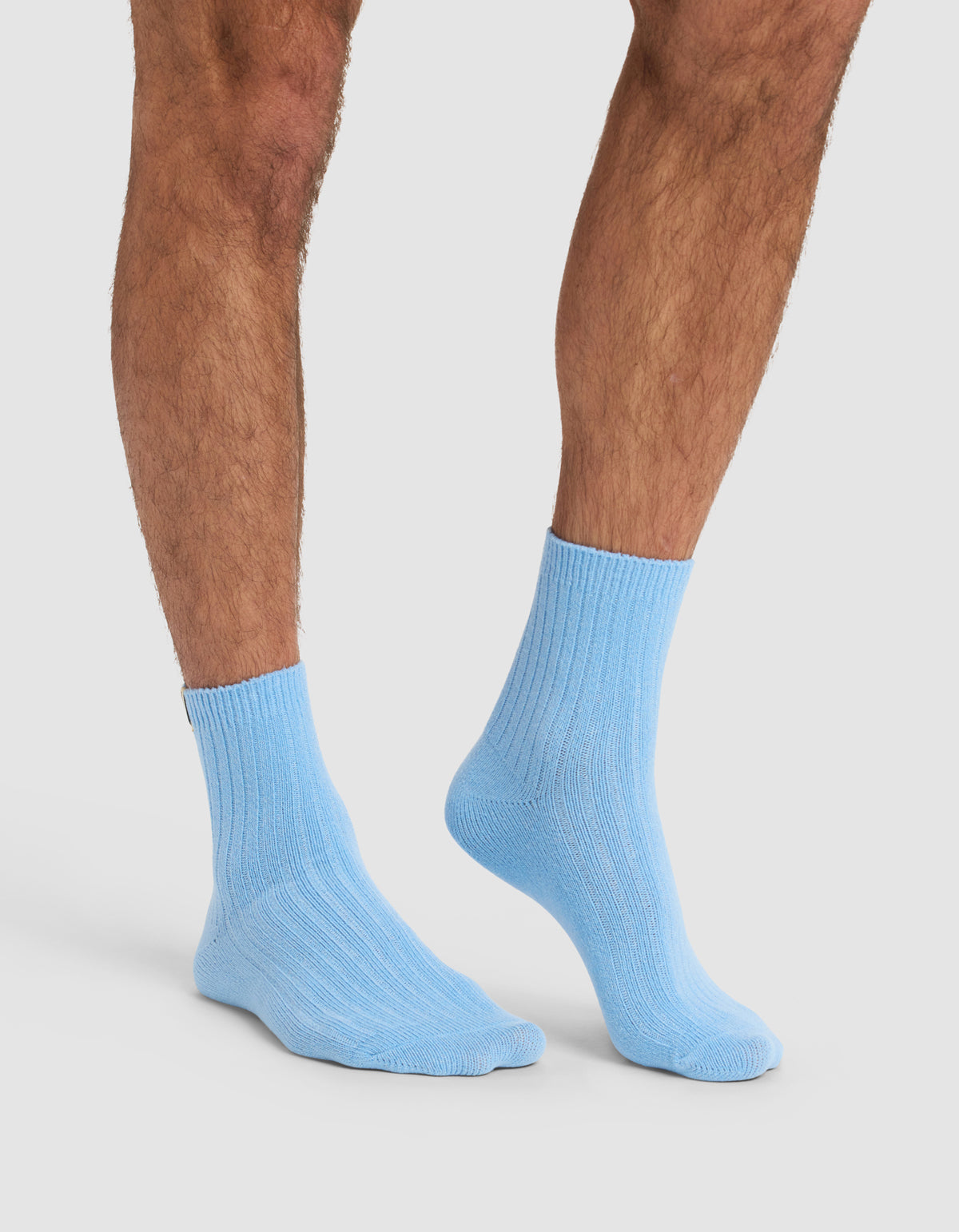A person with hairy legs wears Cozy Earth's The Plush Modern Crew Sock in light blue ribbed style, standing on a white background.