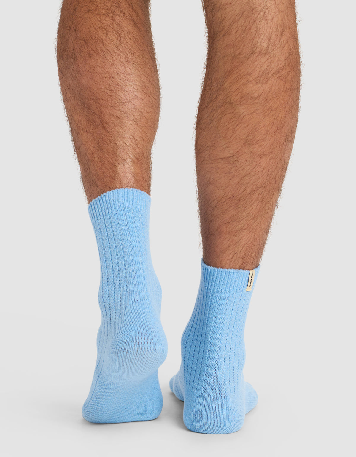 A close-up of a person's lower legs and feet from behind, wearing Cozy Earth’s The Plush Modern Crew Sock in light blue, set against a plain white background.