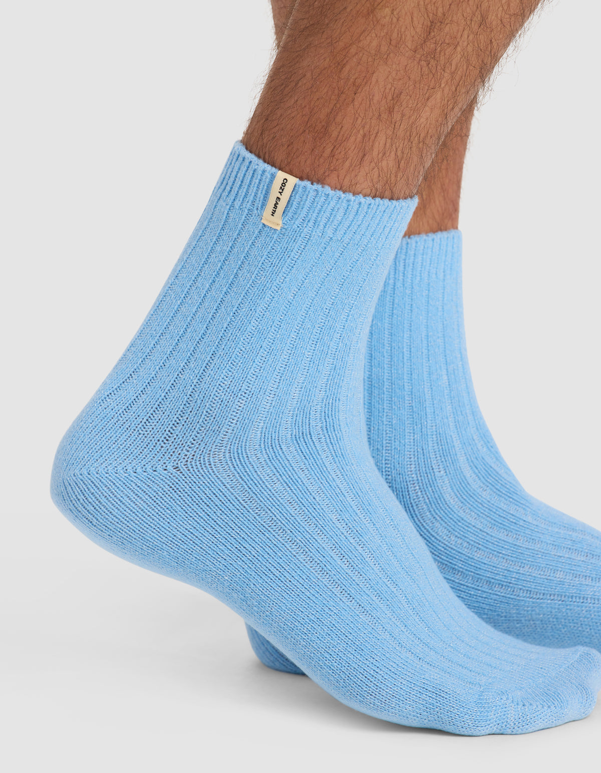 A close-up of a person's lower legs wearing Cozy Earth's The Plush Modern Crew Sock in light blue with a small beige tag near the top edge, standing on a white background.