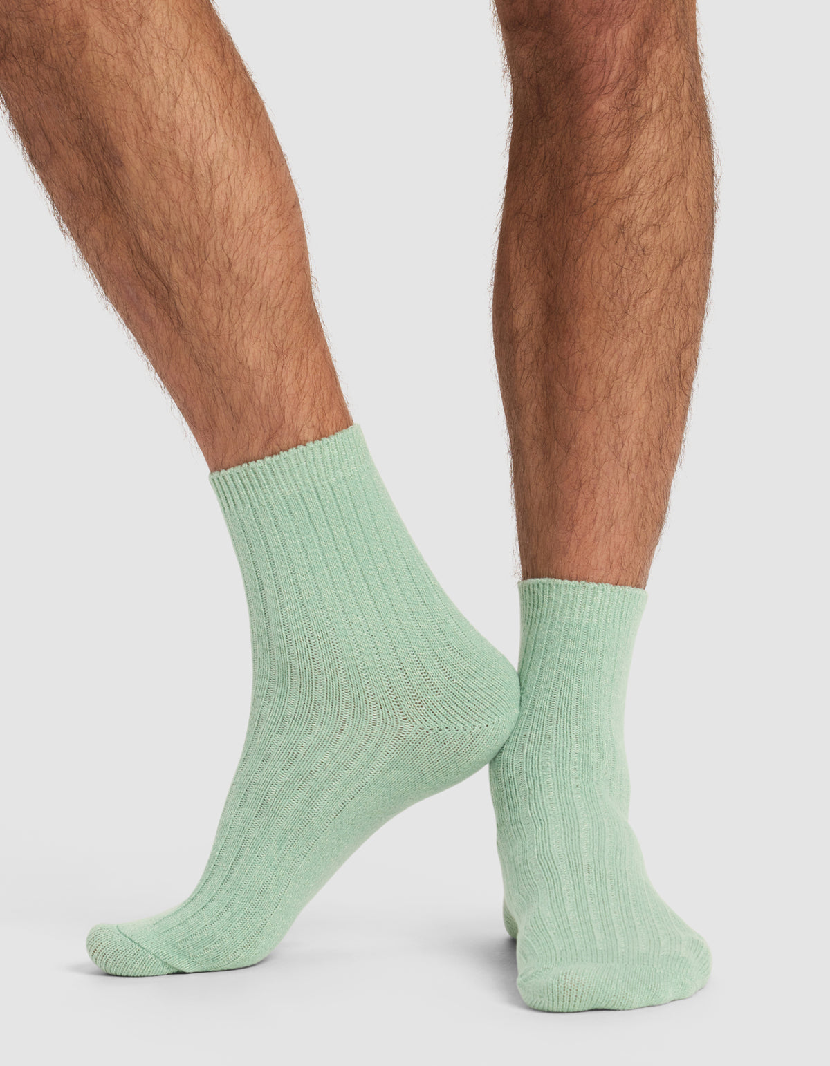 A person with hairy legs is wearing Cozy Earth’s The Plush Modern Crew Sock in light green, standing with one foot slightly crossed behind the other on a plain white background.
