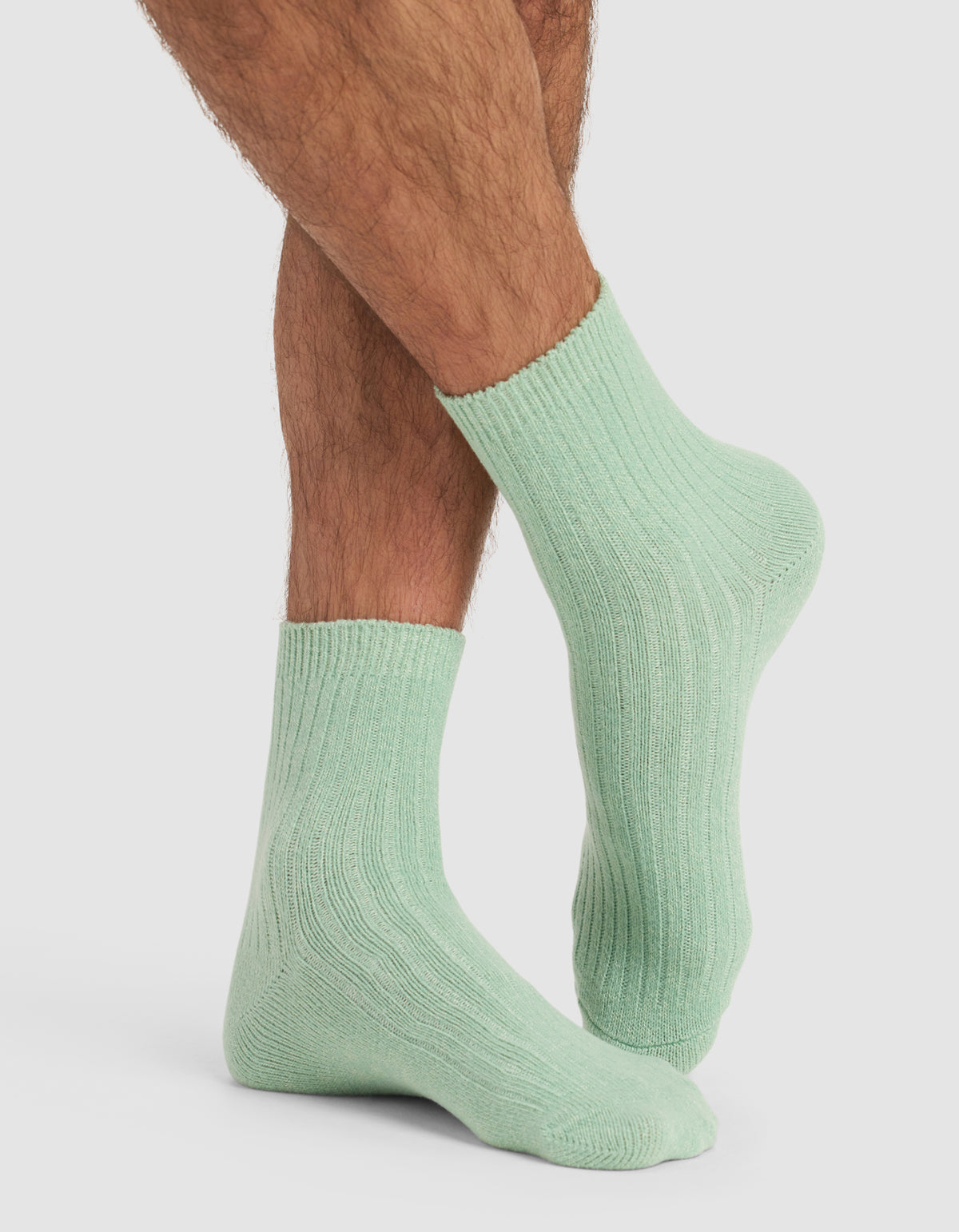 A person with crossed legs is wearing The Plush Modern Crew Sock by Cozy Earth in light green, shown from the calves down against a plain white background.