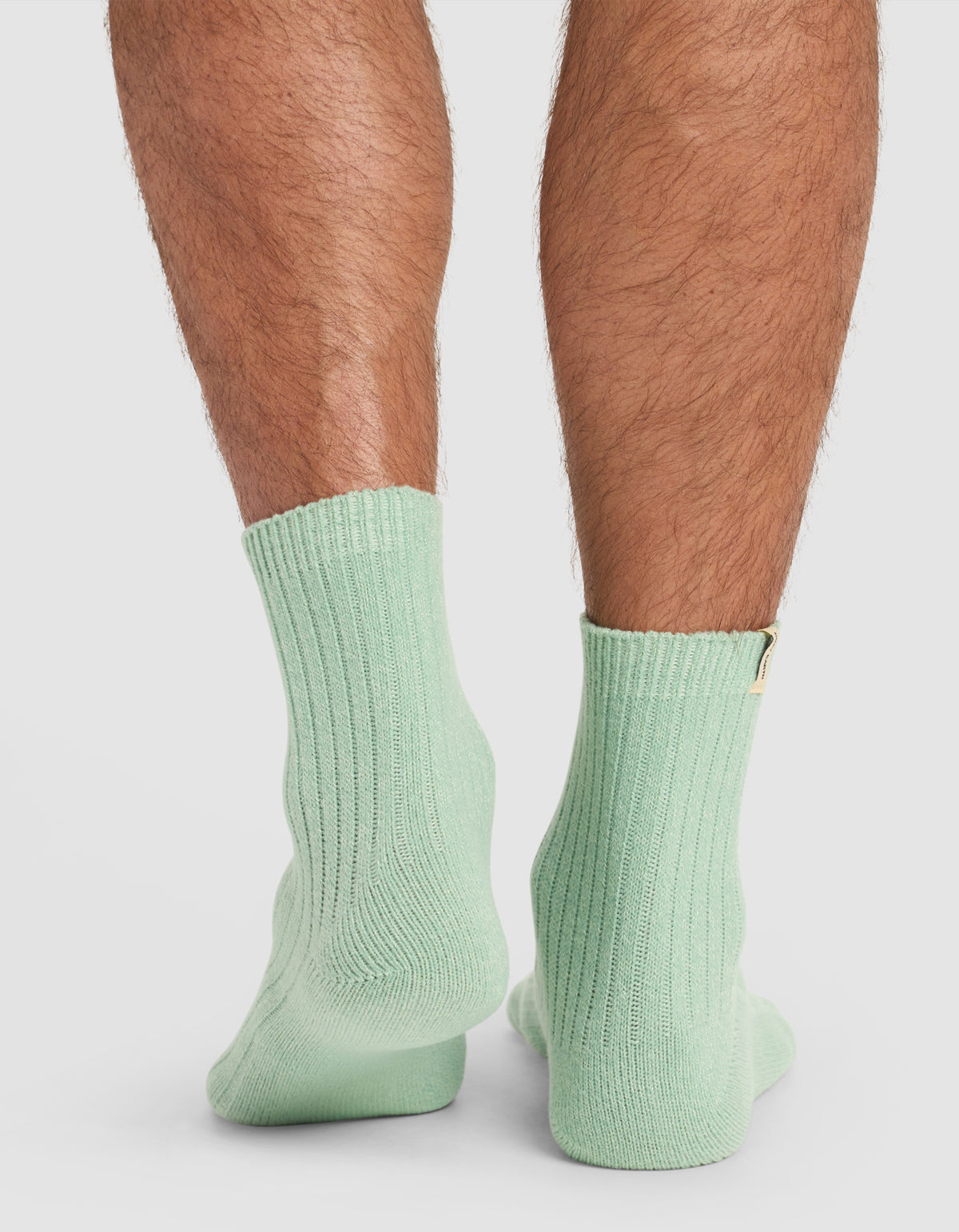 The image displays the back view of a person's lower legs wearing Cozy Earth's The Plush Modern Crew Sock in light green, standing on a white surface.