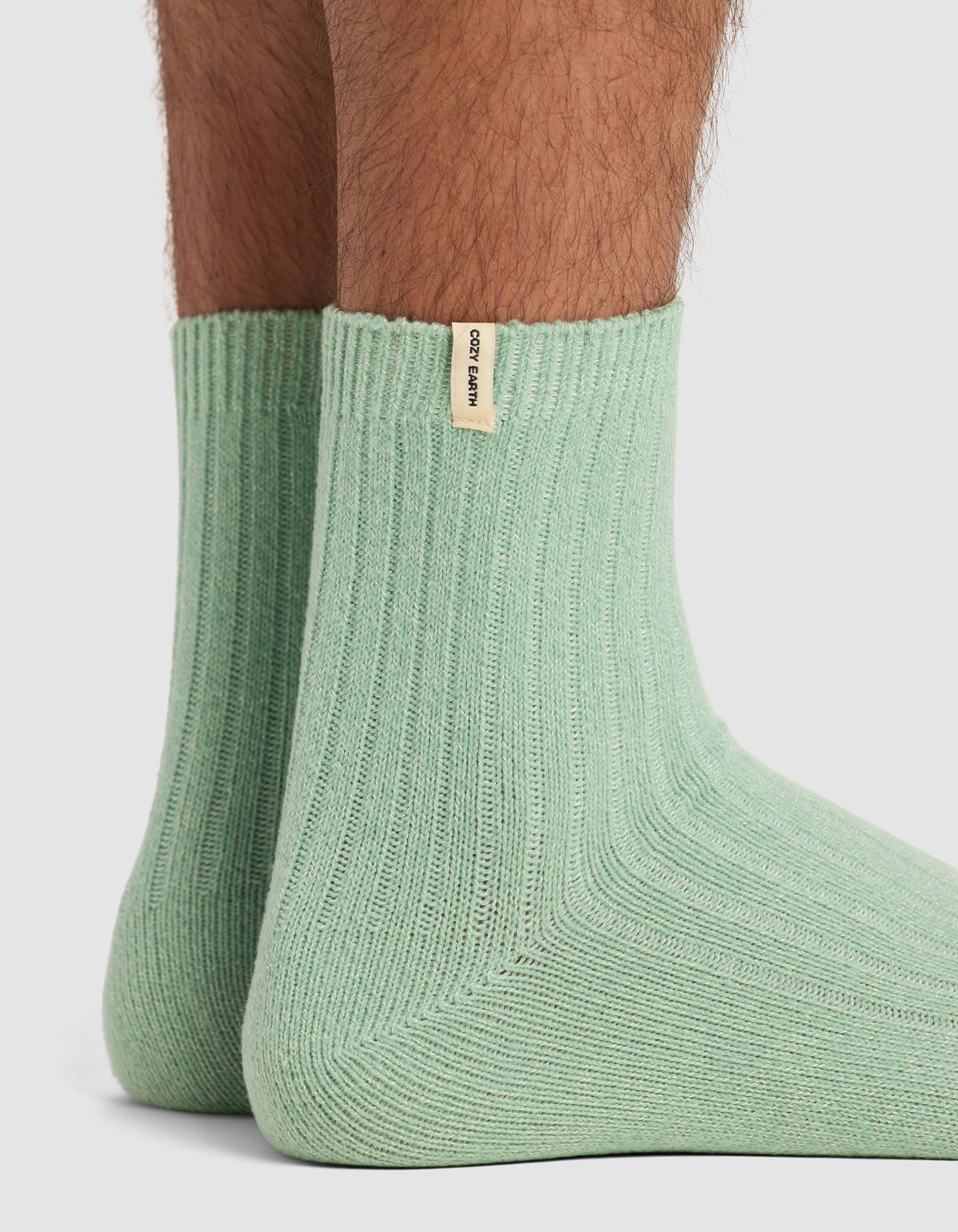 Close-up of a person's lower legs and feet wearing The Plush Modern Crew Sock by Cozy Earth in light green, standing on a white surface. A small beige tag with black text is attached near the top of one sock.