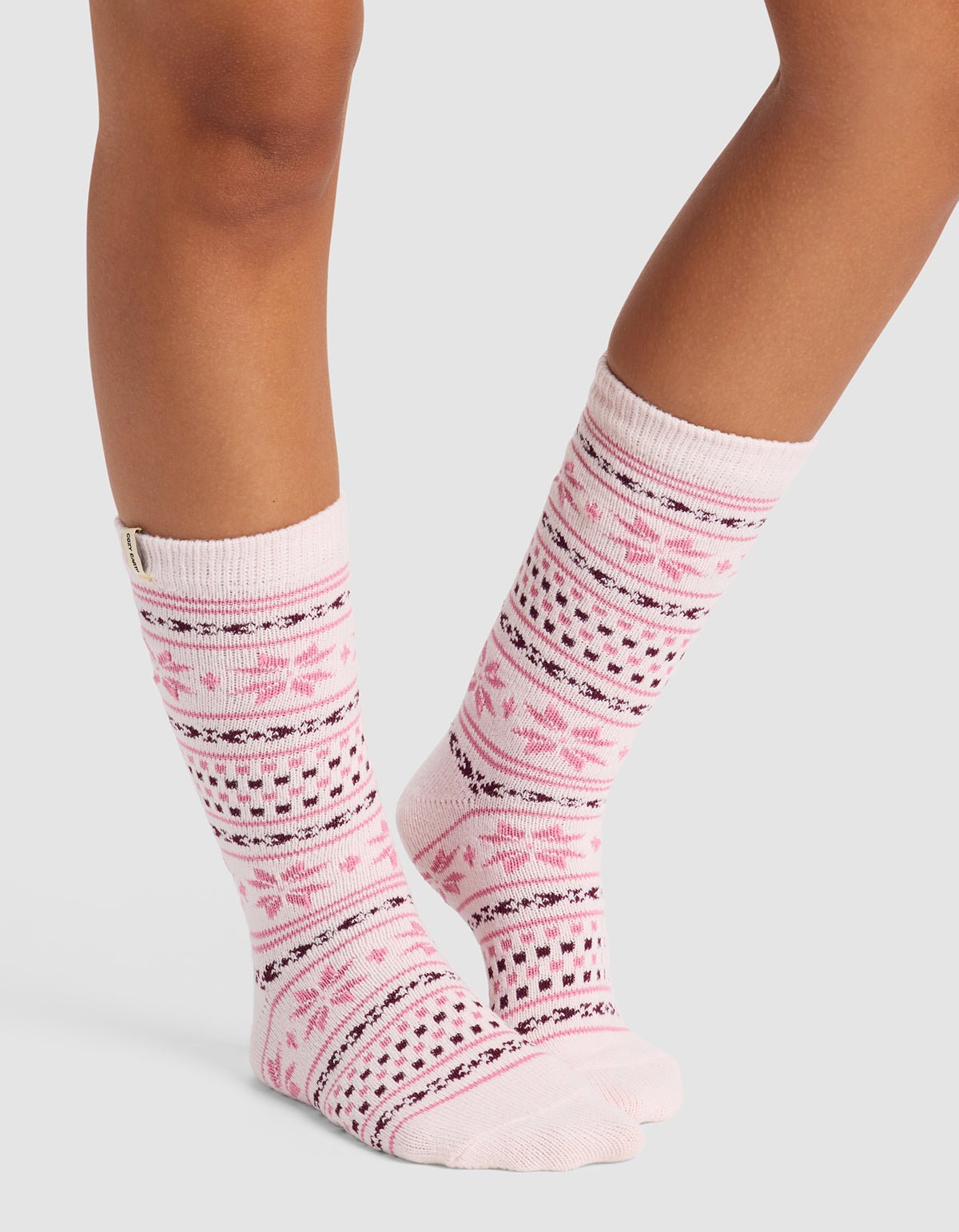 A person stands on a light gray background wearing Cozy Earth's Snowflake Plush Lounge Sock, a knee-high pink and white sock with Nordic snowflakes and geometric patterns.
