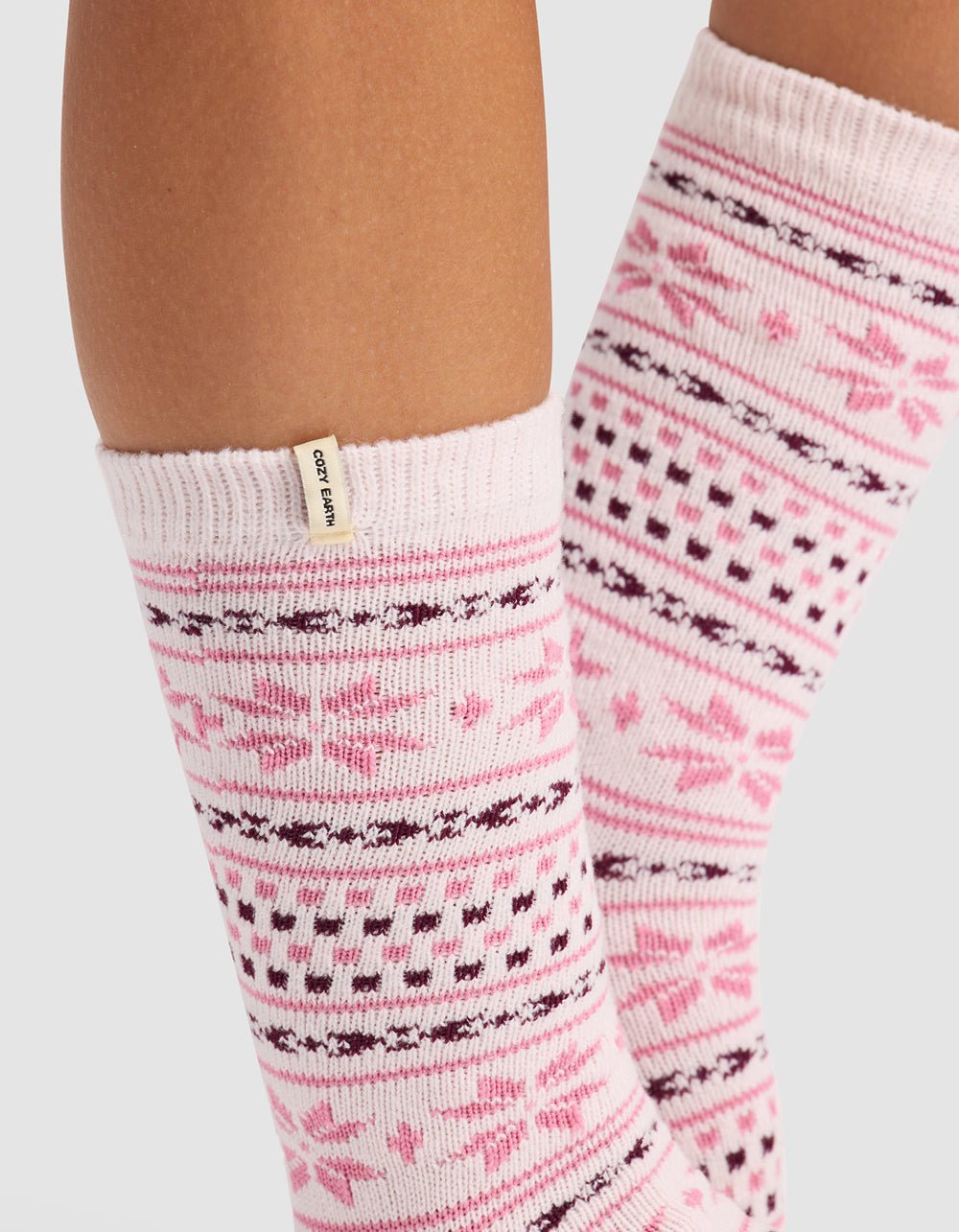 Close-up of a person's lower legs wearing Cozy Earth's The Snowflake Plush Lounge Sock in light pink with dark pink and burgundy snowflake and geometric patterns, featuring a small beige tag on the sock cuff.