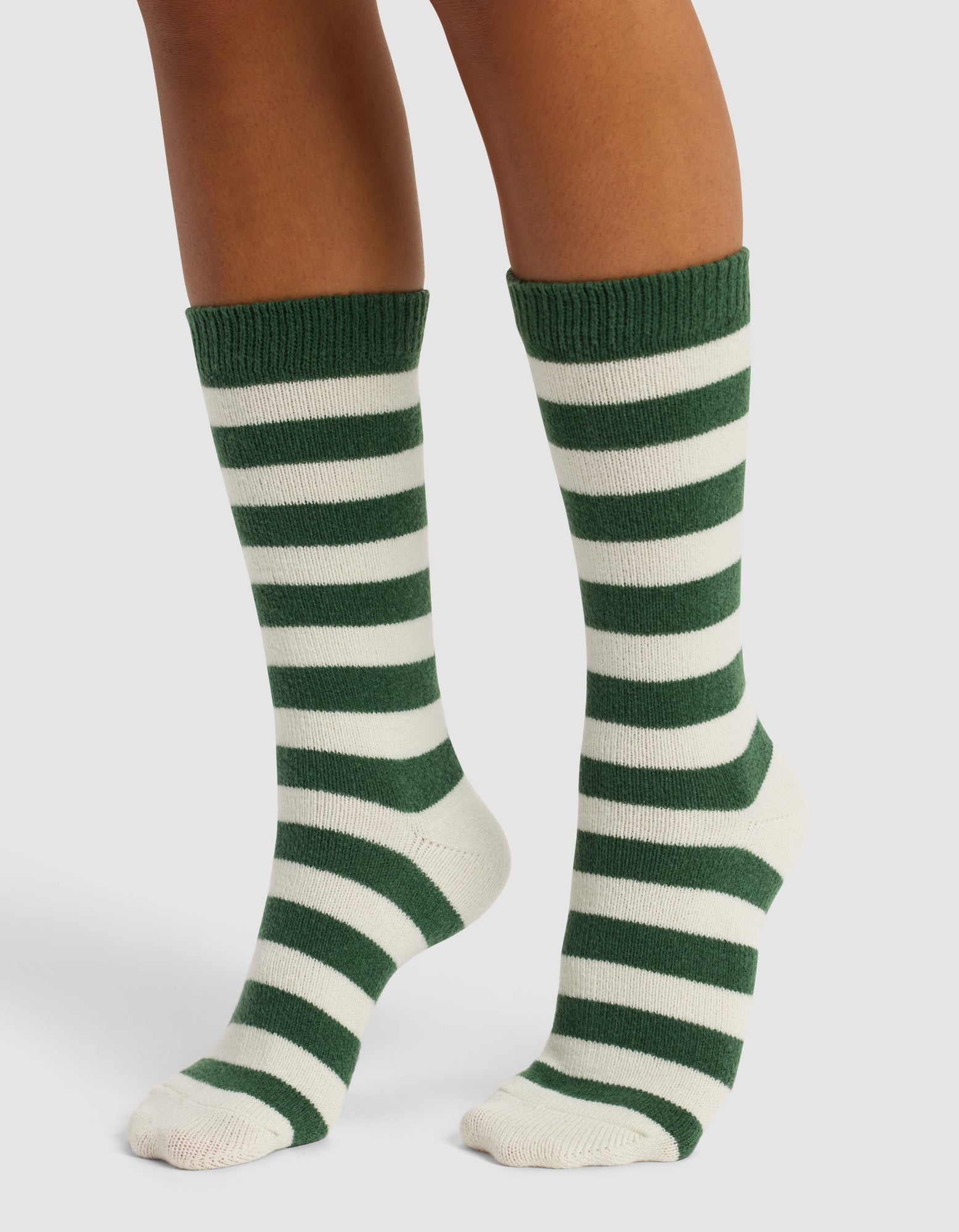 A pair of feet wear Cozy Earth's The Striped Plush Lounge Sock—knee-high, with horizontal dark green and white stripes—standing on a white background.
