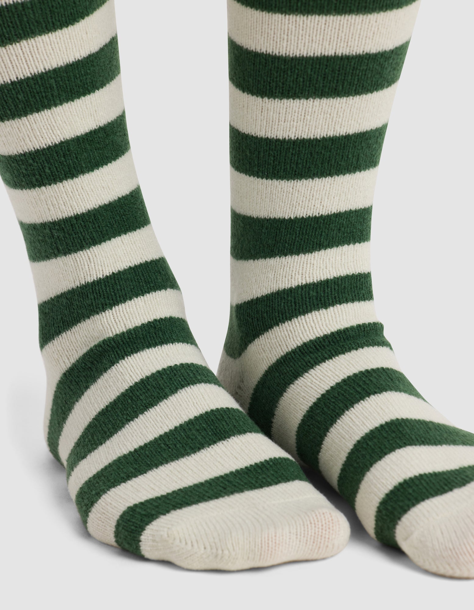 Close-up of feet wearing Cozy Earth's The Striped Plush Lounge Sock, featuring thick, horizontal dark green stripes on white, standing on a white background.