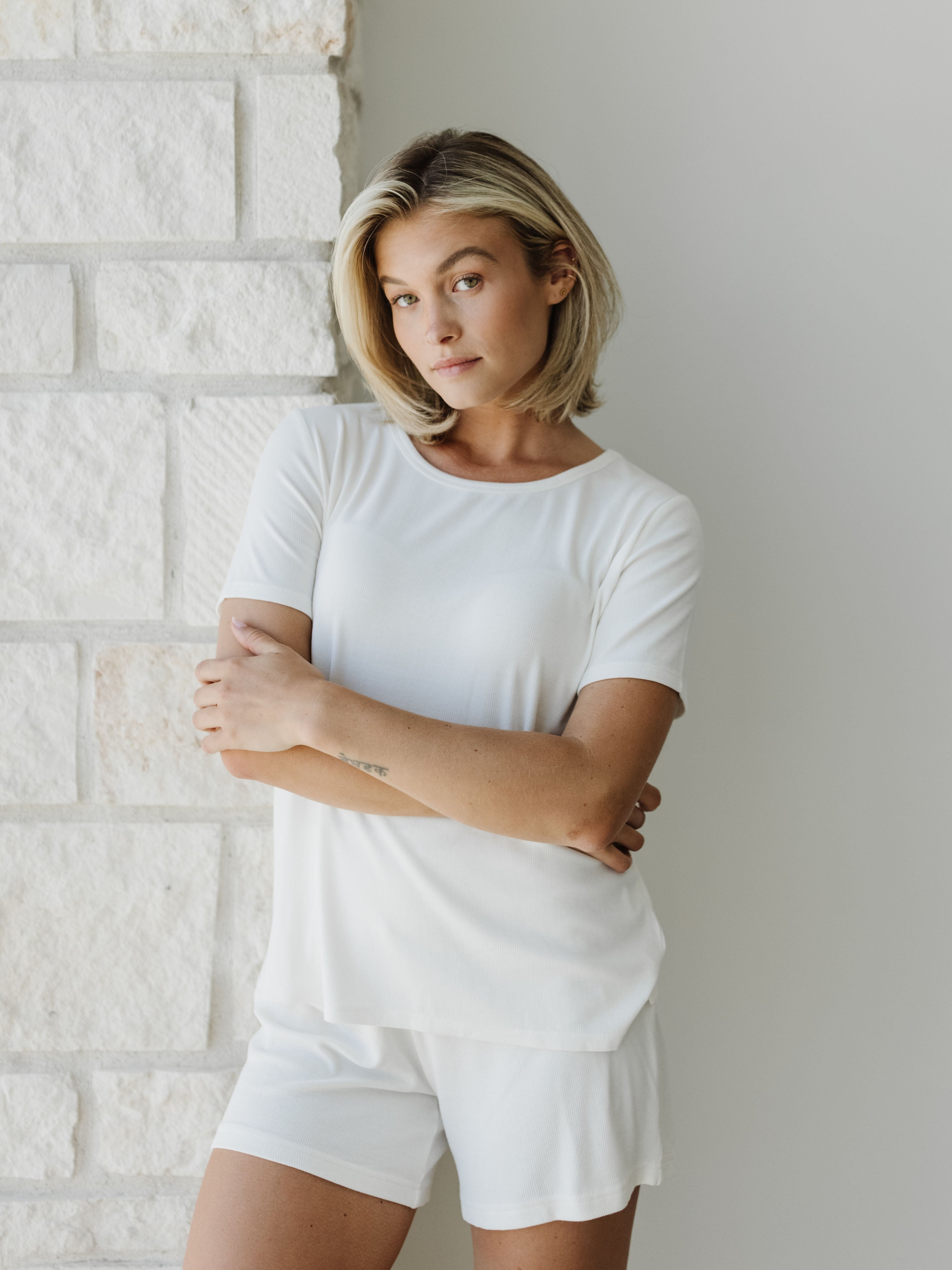 Woman wearing a white Cozy Earth Rib Knit Short Sleeve Lounge Top and white shorts against a white stone background |Color:Ivory