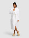 A woman stands against a plain background wearing the Cozy Earth Waffle Bath Robe tied at the waist and beige slippers. Her hair is tied back, with one hand near her collar and one at her side.|Color:White