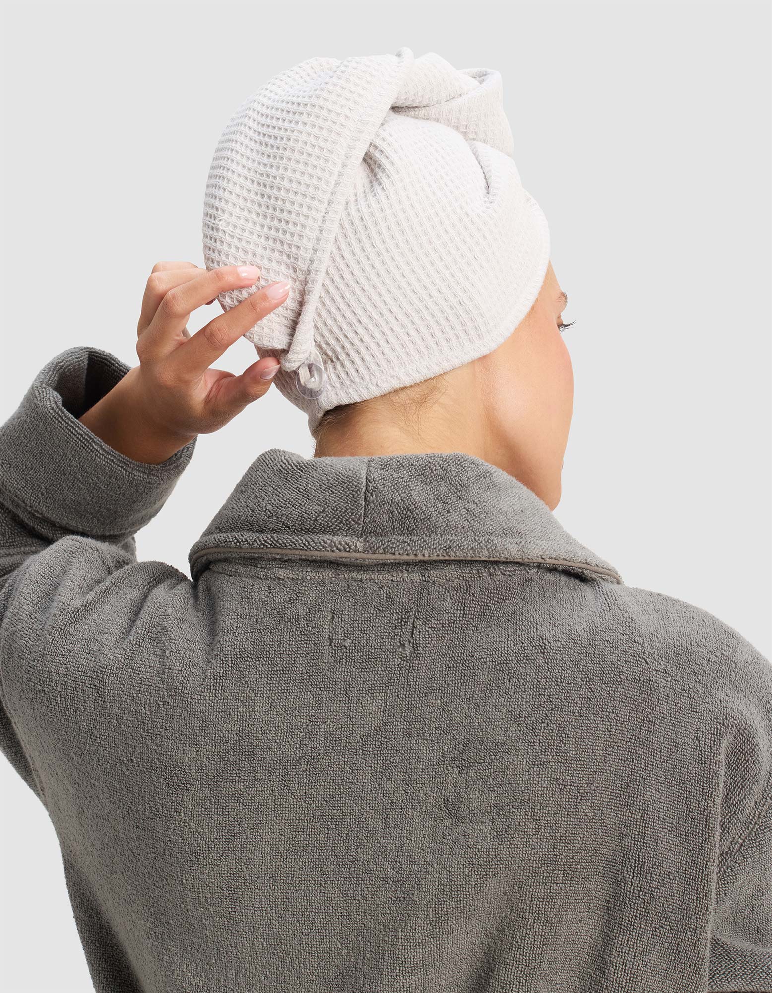 A person in a gray bathrobe is seen from behind, adjusting a Cozy Earth Waffle Hair Towel wrapped around their head. The background is plain and light-colored.