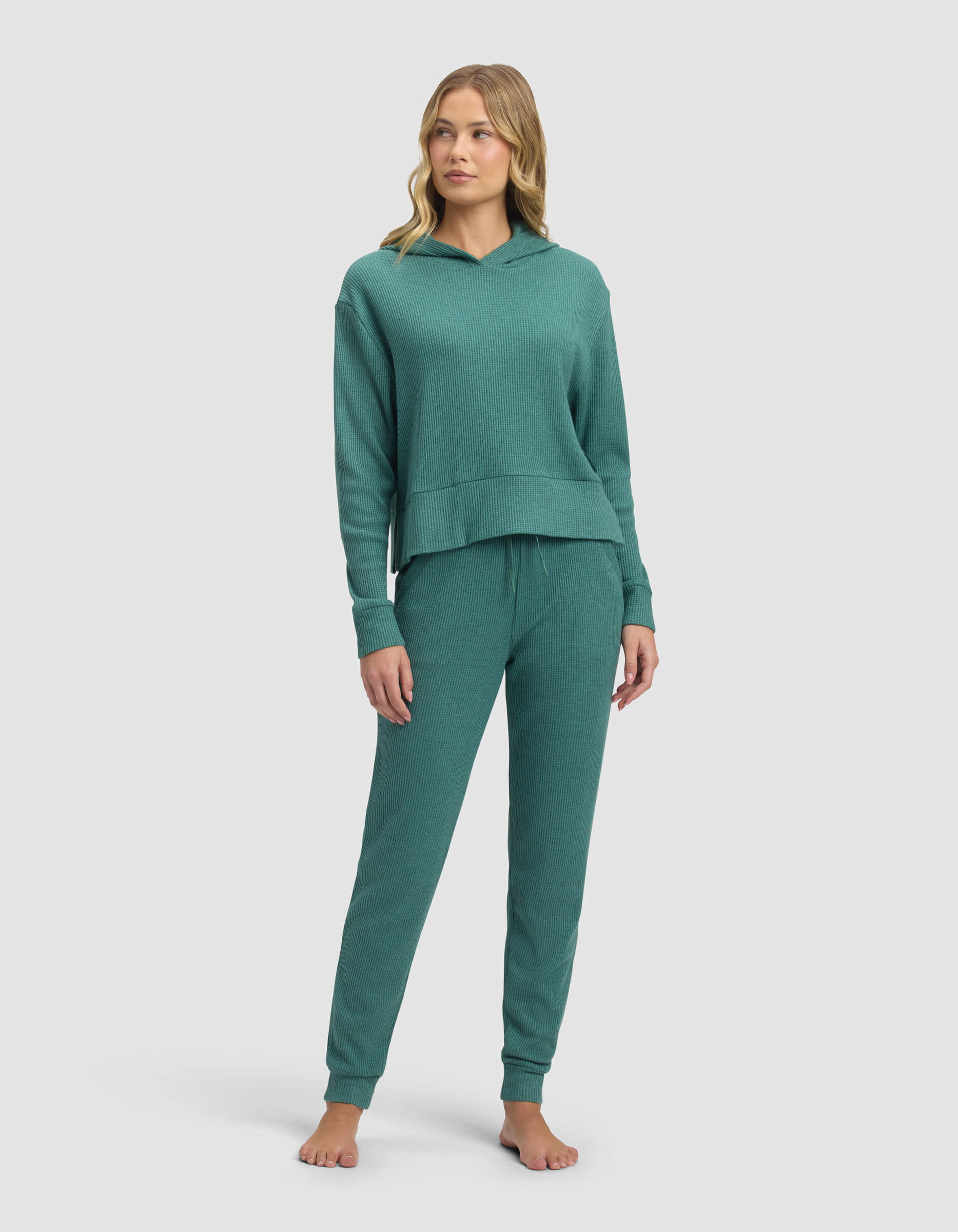 A woman stands barefoot in the Cozy Earth Women's Blake Waffle Hoodie & Jogger Set, featuring a matching teal long-sleeve hoodie and drawstring pants, against a plain light background.
