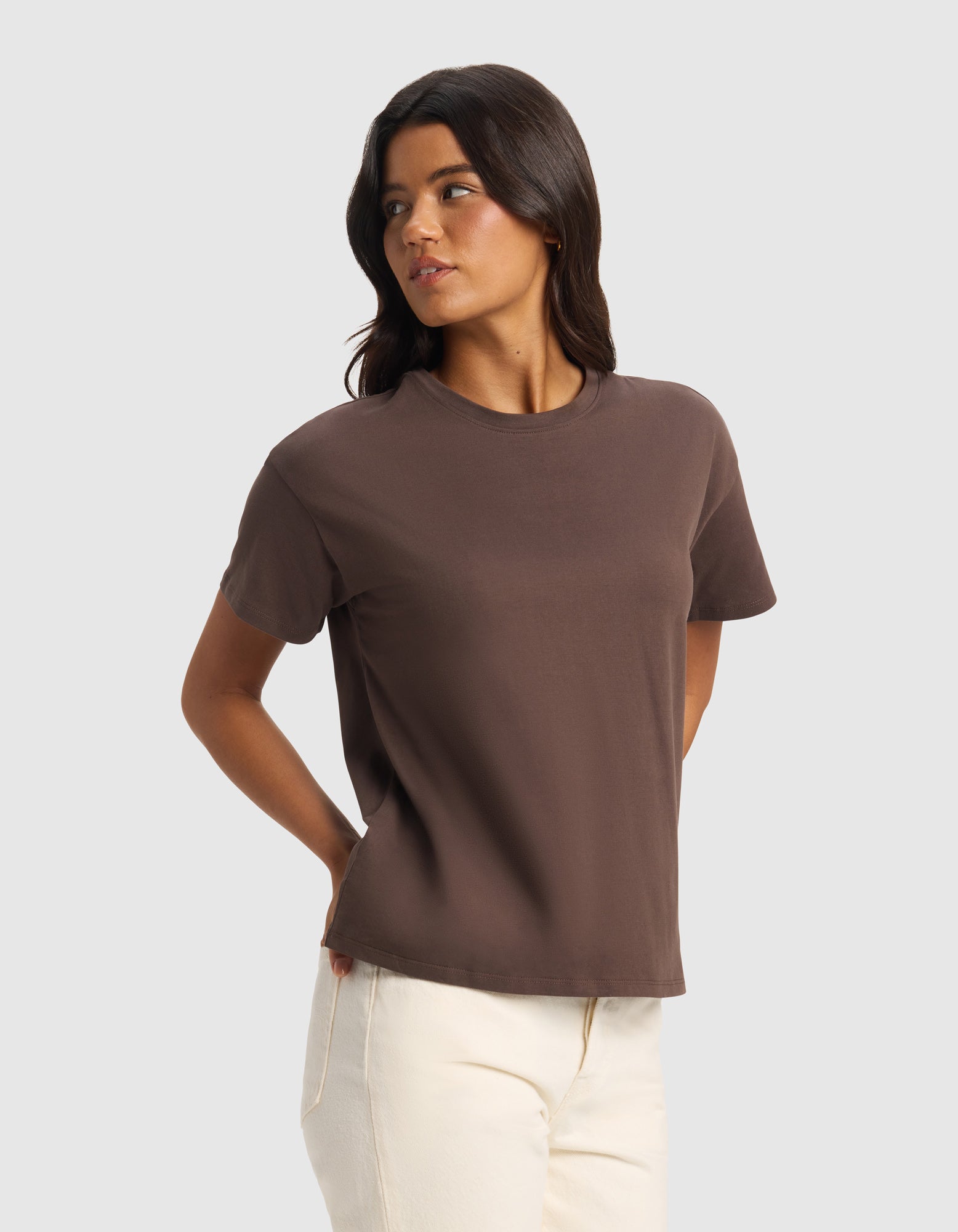 A woman with long dark hair wears Cozy Earth's Women's All Day Tee in brown and off-white pants, standing against a light gray background with her hands behind her back, looking to her left.|Color:Cacao