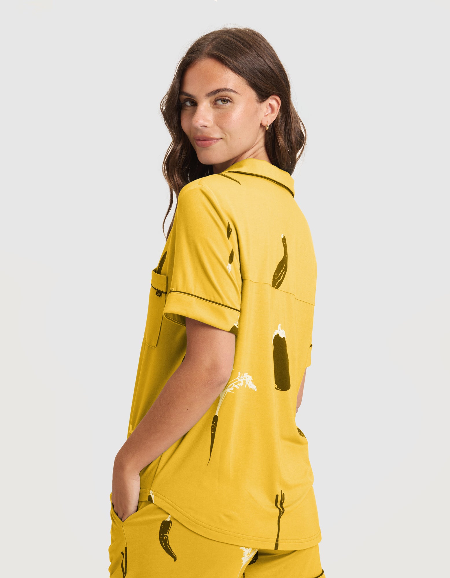 A woman with long brown hair models the Women's Bamboo Stretch-Knit Short Sleeve Pajama Top (Veggie) and matching shorts, featuring a veggie print, as she stands sideways against a plain light background.