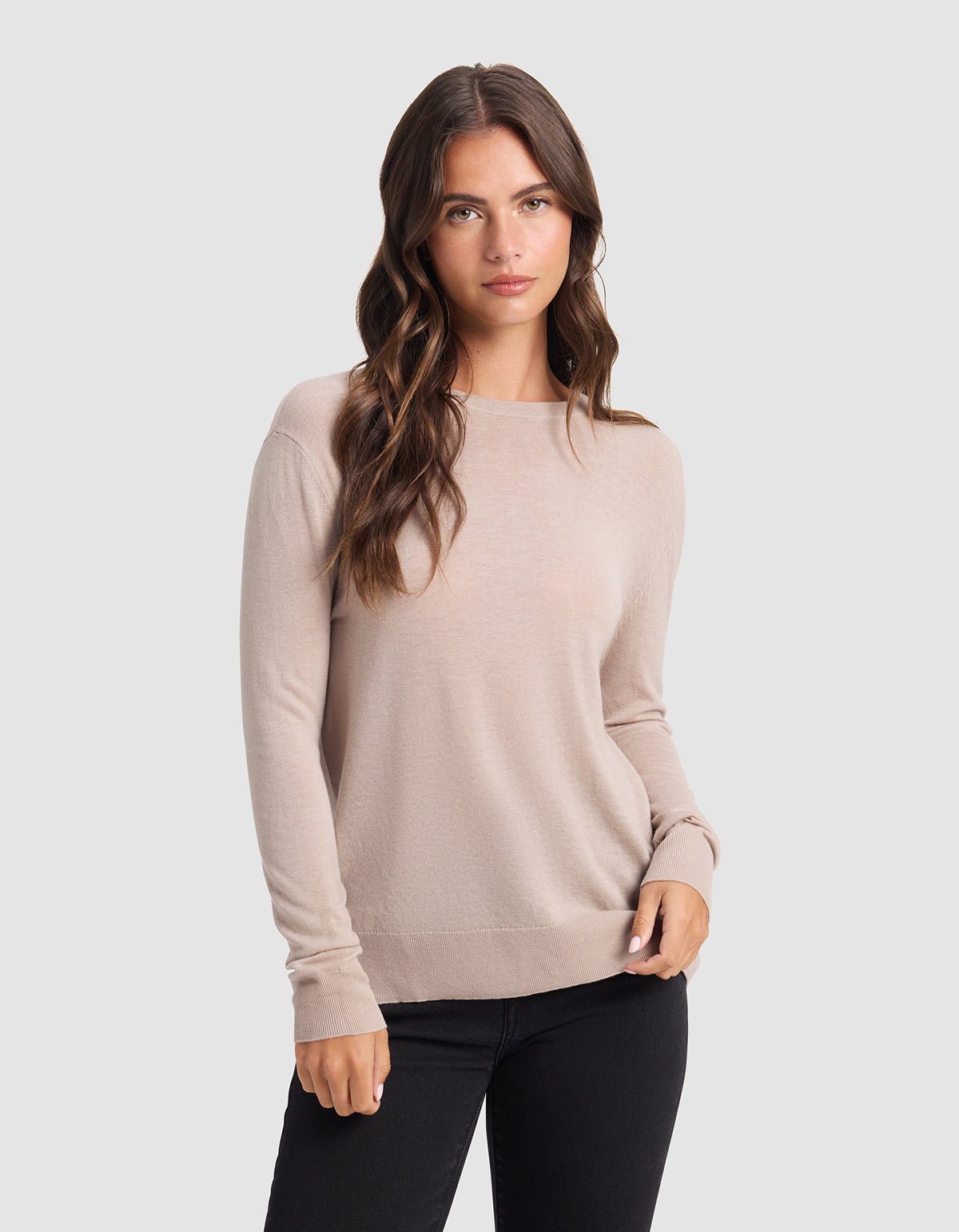 A woman with long wavy brown hair wears the Cozy Earth Women's AirKnit Crewneck Sweater in light beige and black pants, standing against a plain light gray background and gazing at the camera with a relaxed expression. |Color:Taupe