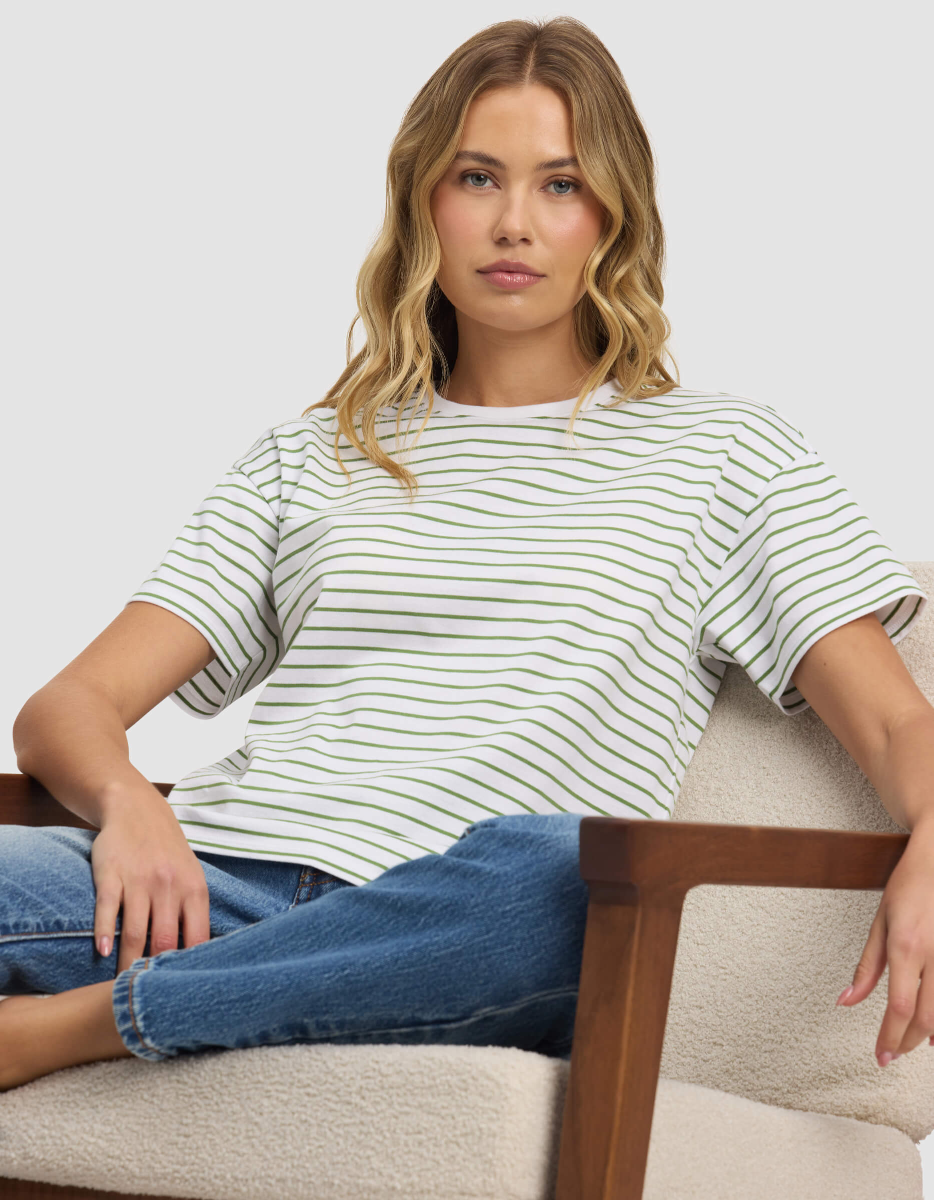 A woman with wavy blonde hair sits on a light-colored armchair, wearing Cozy Earth's Women's All Day Tee and blue jeans, looking directly at the camera with a neutral expression.|Color:Clover Stripe