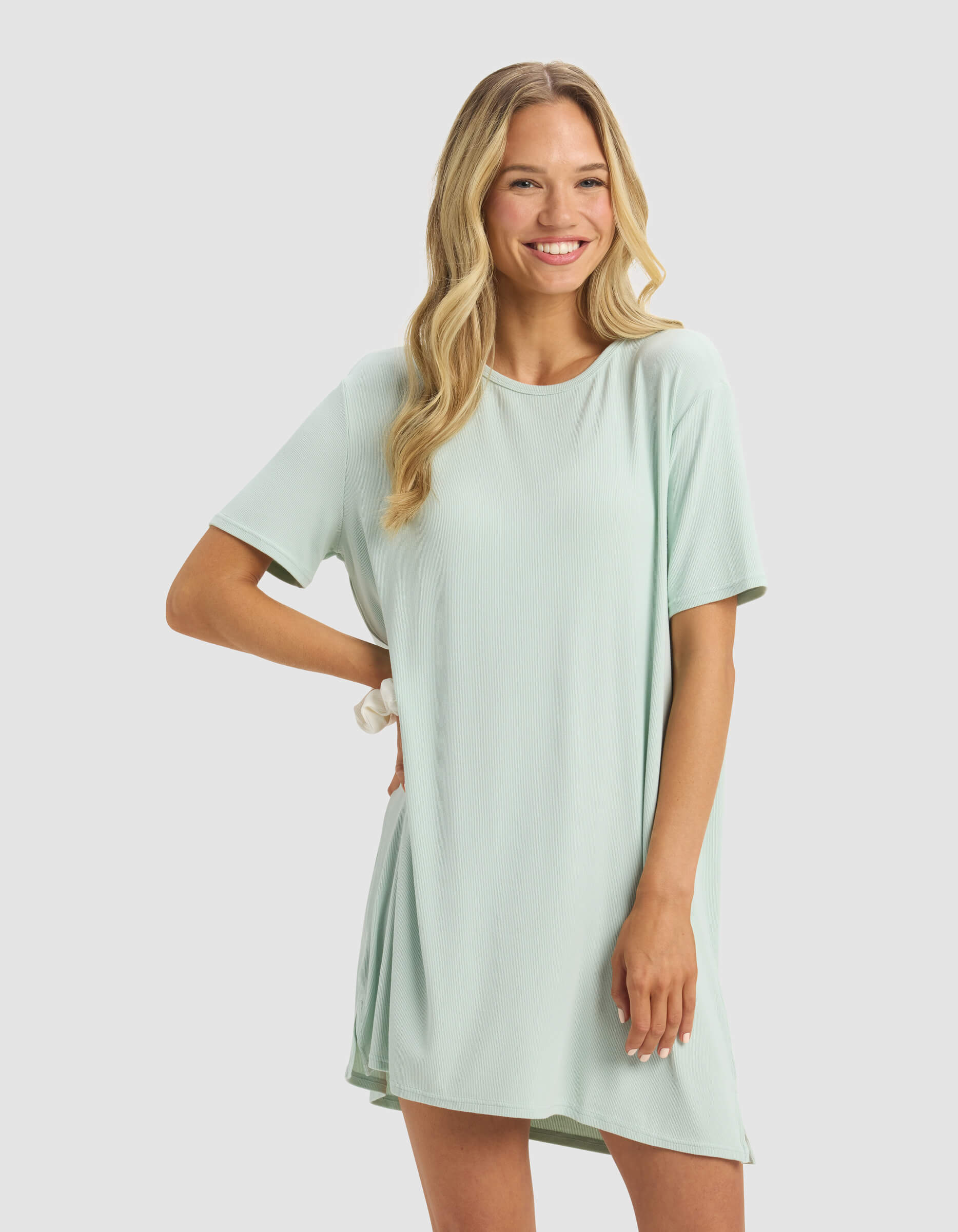 A smiling woman with long blonde hair poses with one hand on her hip against a light gray background, wearing the Cozy Earth Women's Bamboo Rib-Knit Boyfriend Dress in light green.|Color:Seafoam
