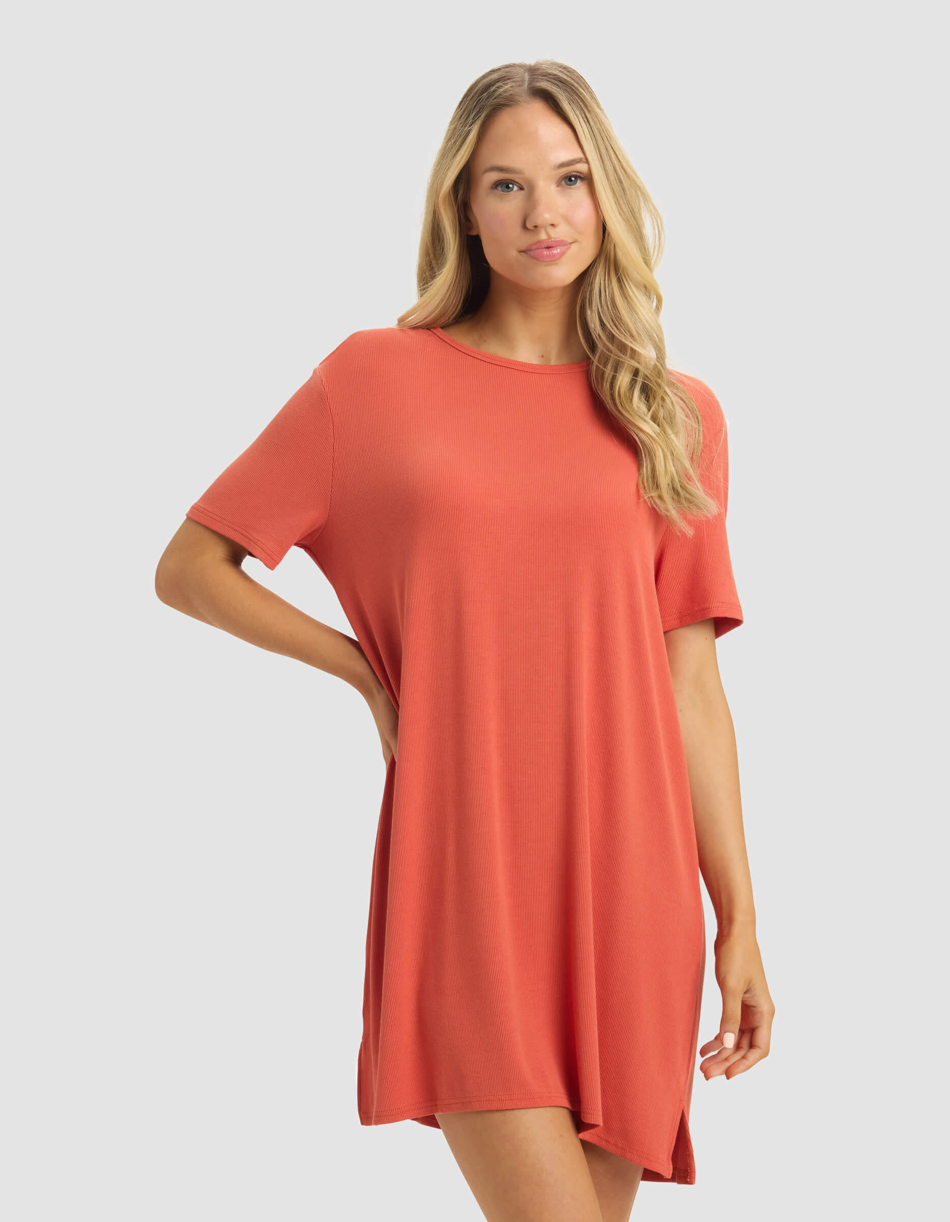 A woman with long blonde hair wears the Cozy Earth Women's Bamboo Rib-Knit Boyfriend Dress in coral-orange, standing against a light gray background with one hand on her hip and a neutral expression.|Color:Sunset