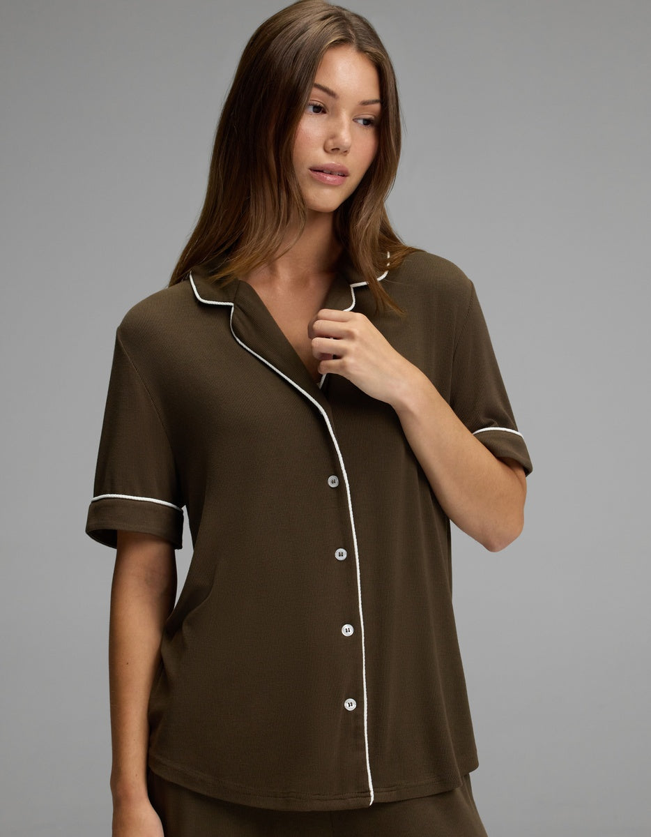 A woman with long brown hair wears the Cozy Earth Women’s Bamboo Rib-Knit Classic Short Sleeve Pajama Top, featuring dark brown fabric with white piping and buttons, while standing against a plain gray background.|Color:Espresso