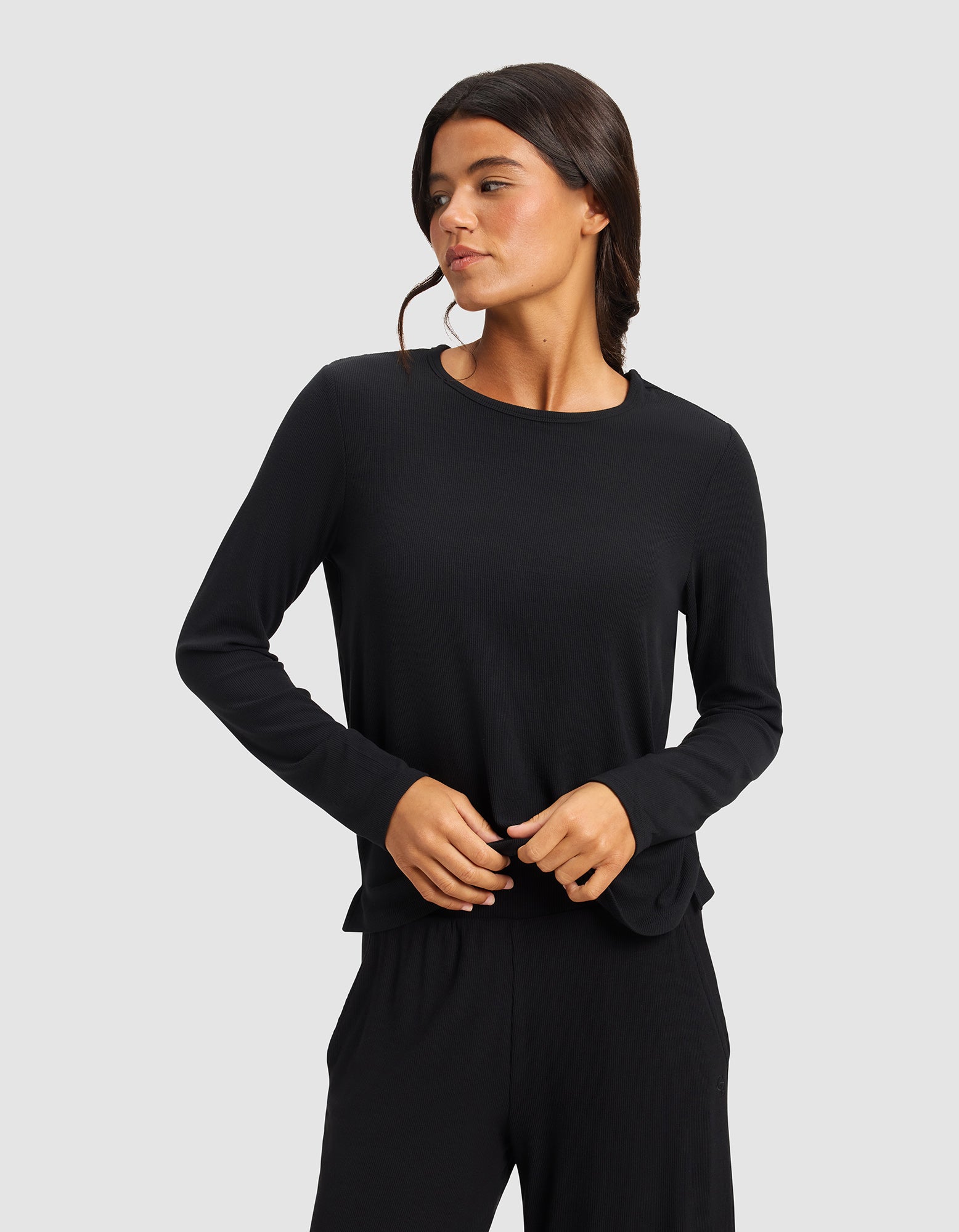 A woman with long dark hair wears the Cozy Earth Women’s Bamboo Rib-Knit Long Sleeve Lounge Top in black, paired with matching black pants, standing against a plain light gray background and looking to her left.