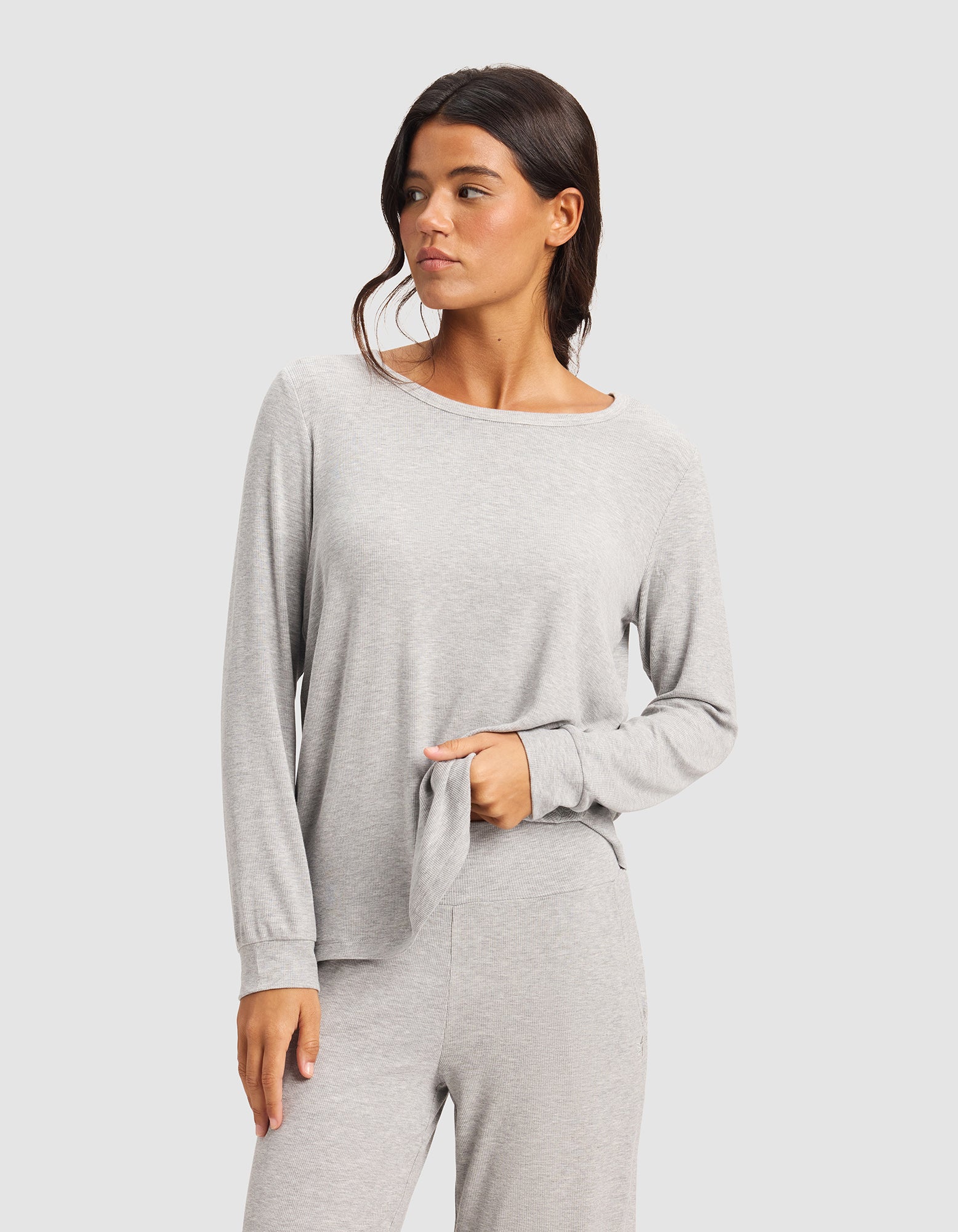 A woman wears the Cozy Earth Women’s Bamboo Rib-Knit Long Sleeve Lounge Top and matching pants, standing against a plain light background, looking left while holding part of her top.|Color:Grey