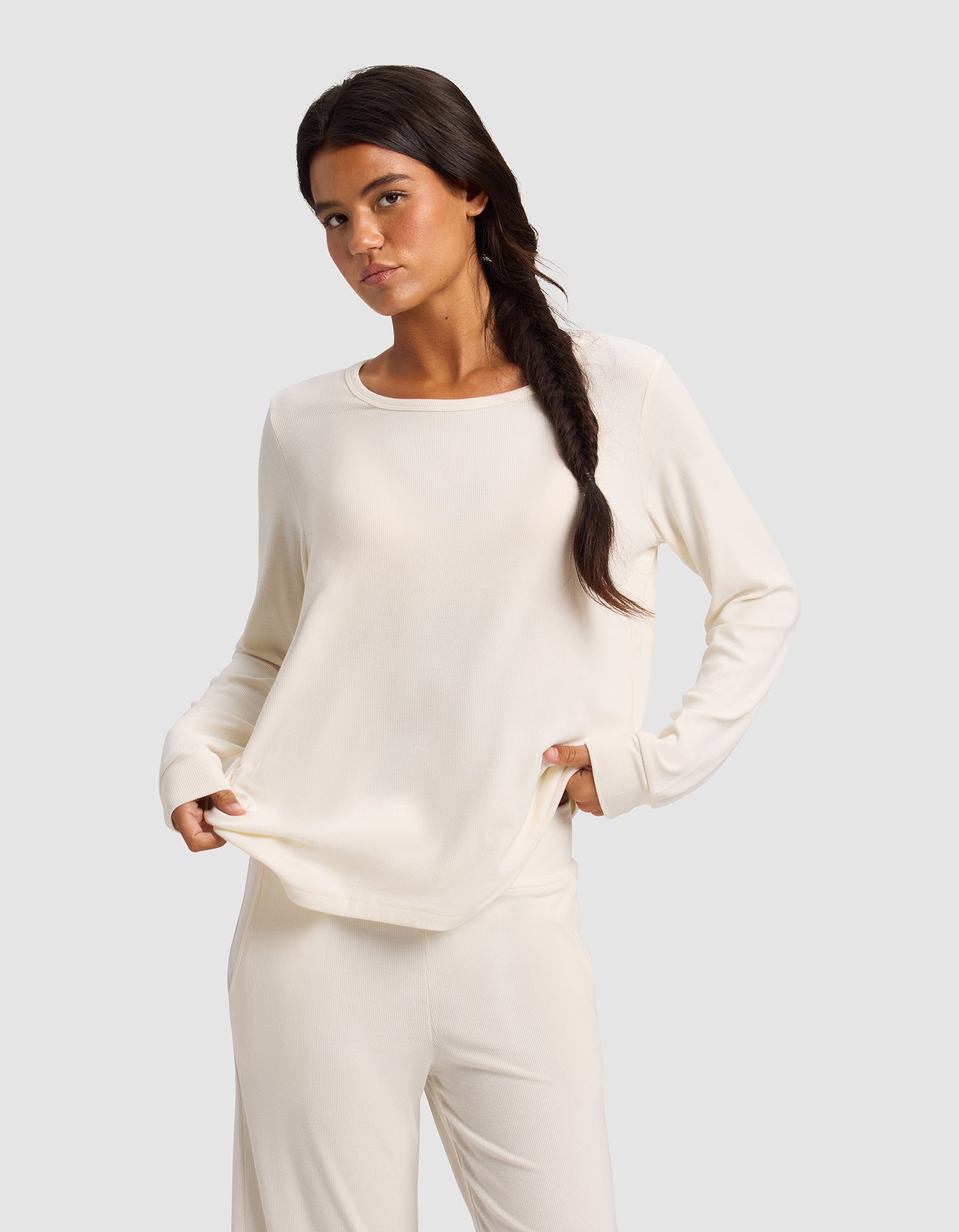 A woman with long dark hair in a loose braid wears the Cozy Earth Women’s Bamboo Rib-Knit Long Sleeve Lounge Top in cream, paired with matching pants, standing against a light gray background and holding the hem of her top.