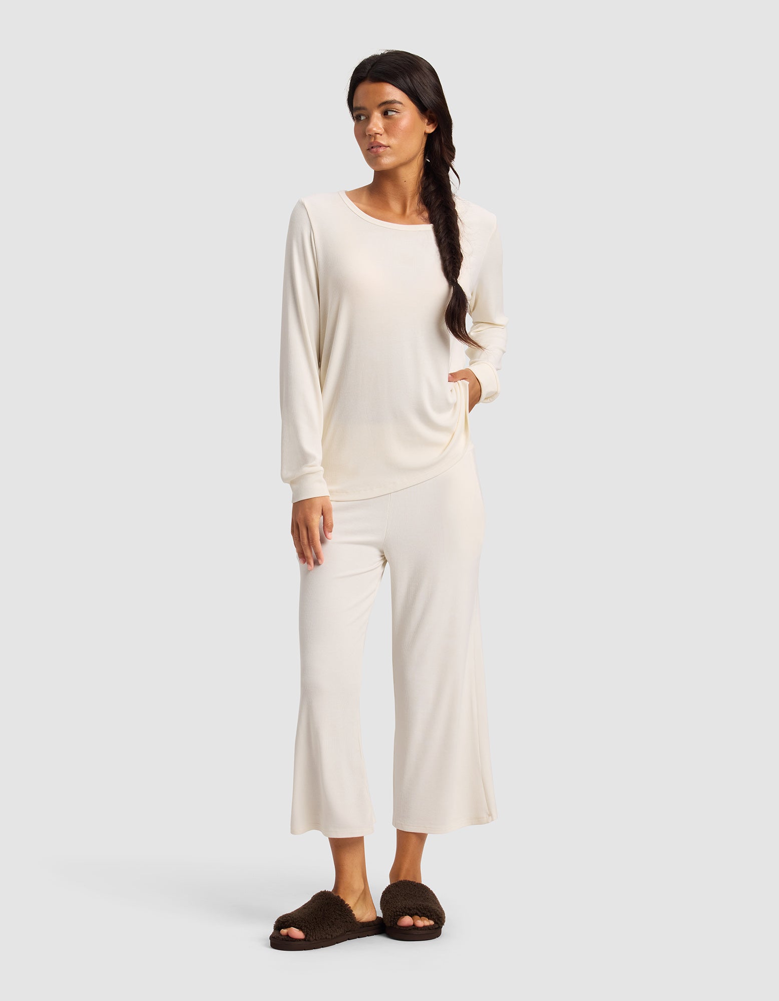 A woman in a Cozy Earth Women’s Bamboo Rib-Knit Long Sleeve Lounge Top and matching wide-leg pants stands with one hand in her pocket. She has long dark hair in a loose braid, wears dark brown slippers, and is set against a light gray background.