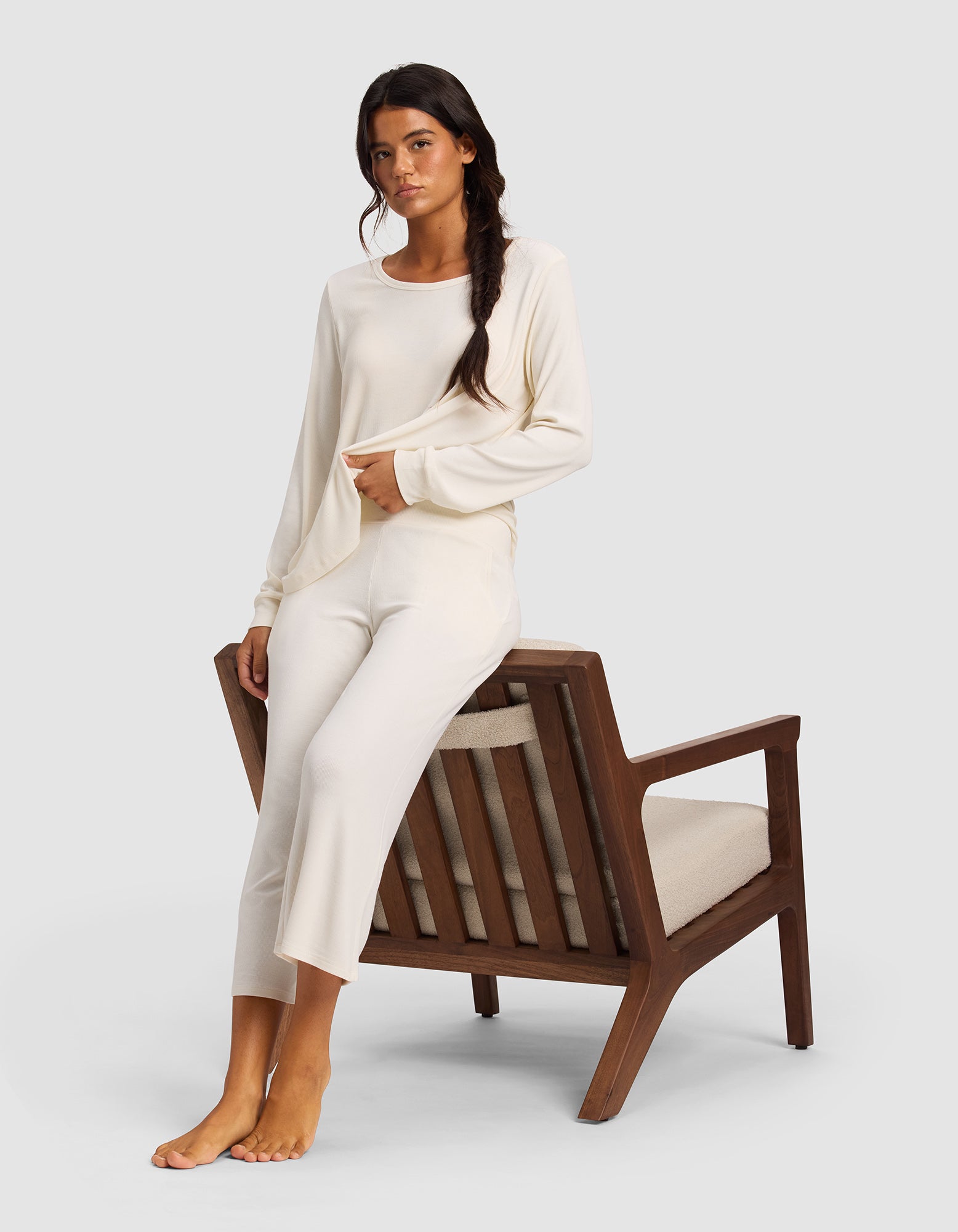 A woman with long dark hair wears the Cozy Earth Women’s Bamboo Rib-Knit Lounge Capri in cream, sitting sideways on a cushioned wooden chair with one foot down and the other bent, against a plain light background.