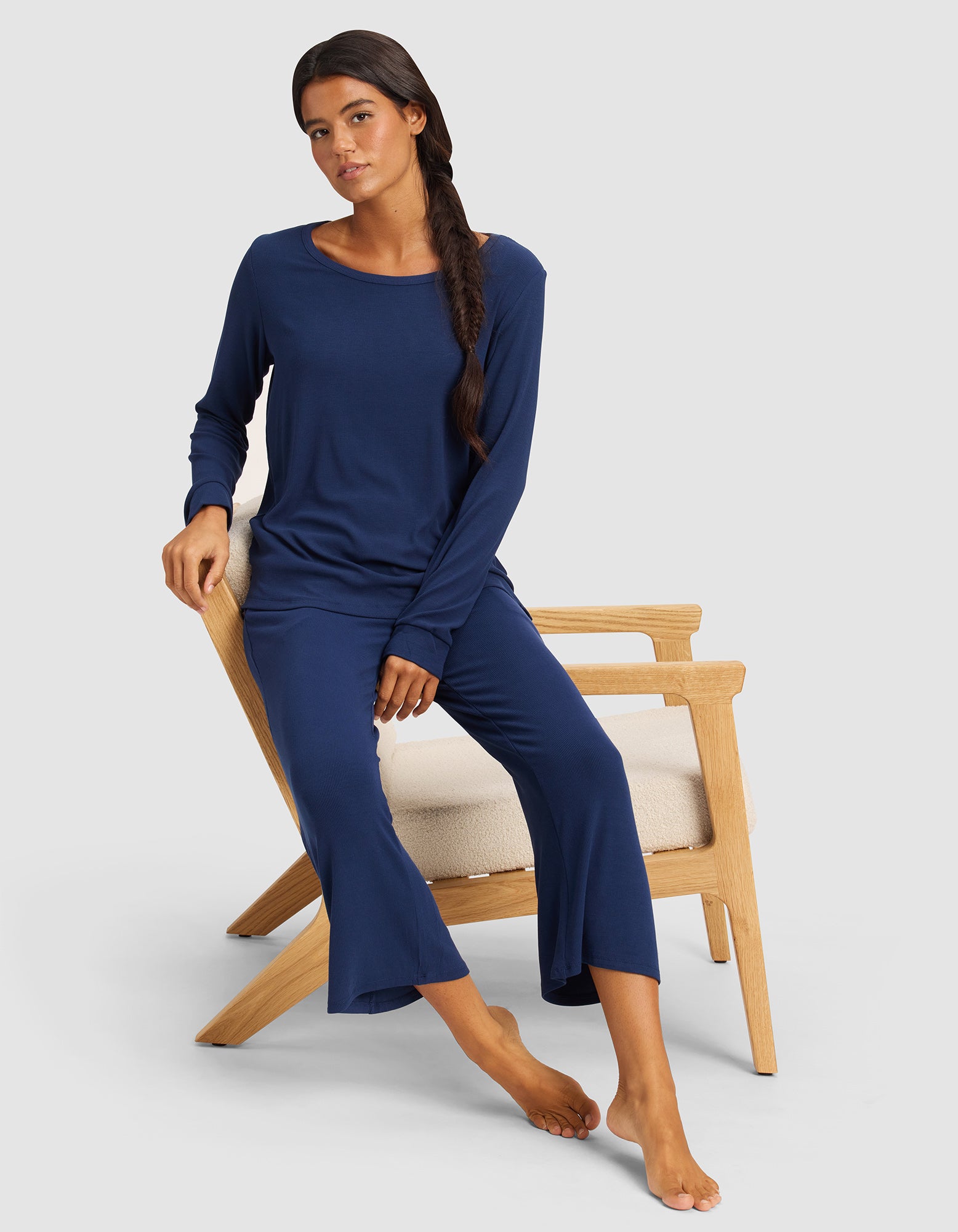 A woman with long dark hair sits barefoot on a wooden chair, wearing Cozy Earth’s Women’s Bamboo Rib-Knit Lounge Capri set in navy blue. She looks relaxed against a plain light background.
