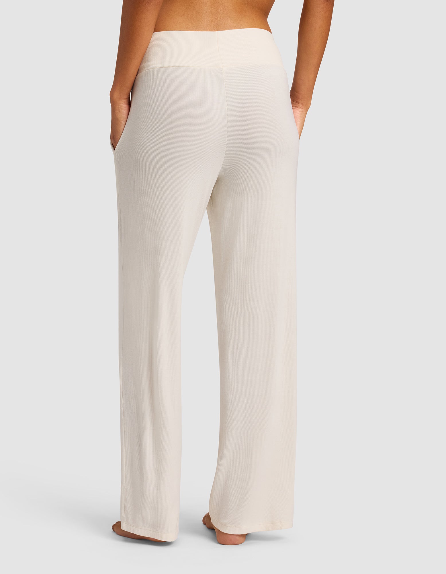 A person wearing Cozy Earth’s Women’s Bamboo Rib-Knit Lounge Pant in light beige, featuring a wide waistband and side pockets, is seen from the back and barefoot against a plain light background.
