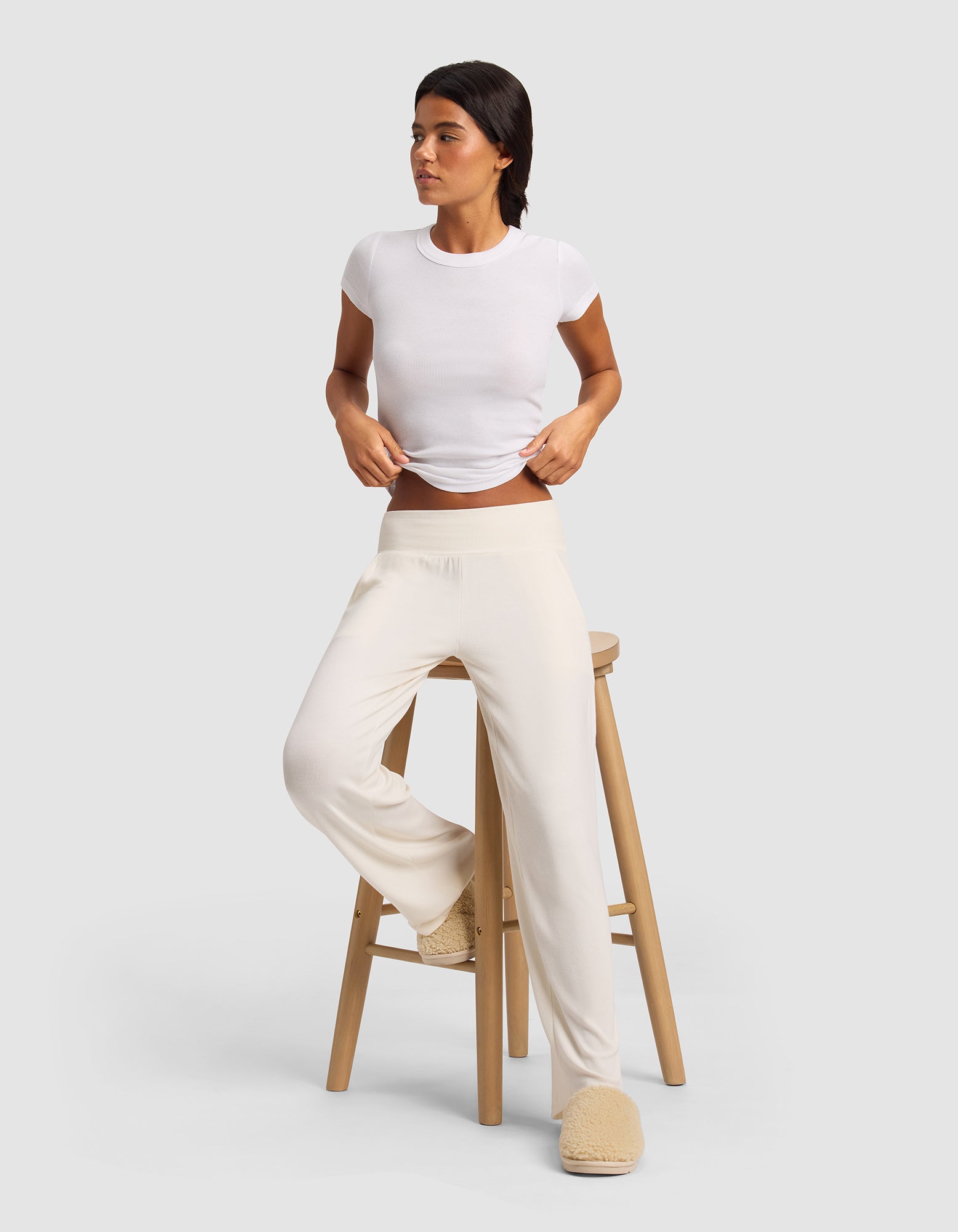 A woman in a fitted white tee, Cozy Earth's Women’s Bamboo Rib-Knit Lounge Pant in cream, and beige slippers leans against a wooden stool, gazing sideways against a plain light gray background.