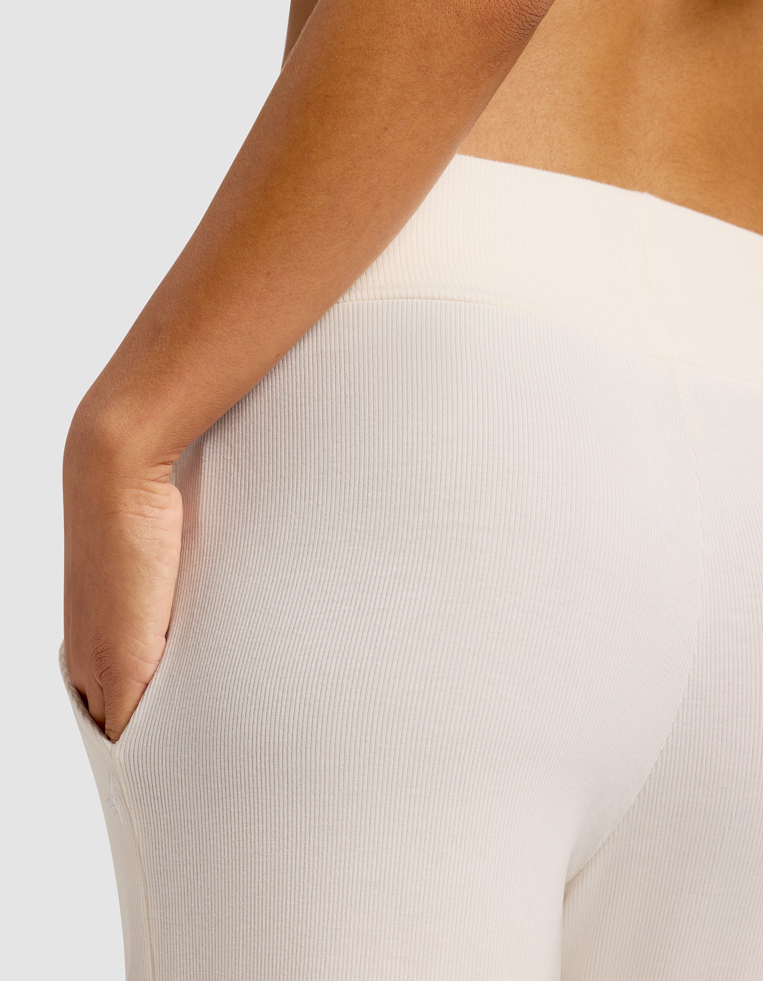 A close-up shows a person wearing Cozy Earth Women’s Bamboo Rib-Knit Lounge Pants in off-white, with their right hand in the side pocket against a plain, neutral background.
