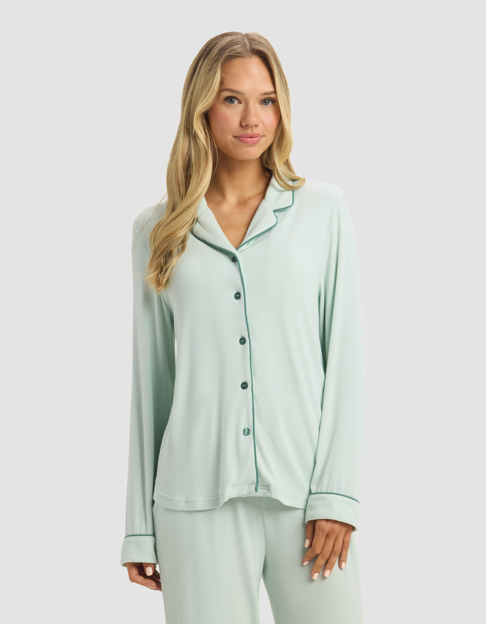 A woman with long blonde hair models Cozy Earth's Women's Bamboo Rib-Knit Classic Long Sleeve Pajama Top in light green with dark green piping, paired with matching pants, against a plain light background.|Color:Seafoam
