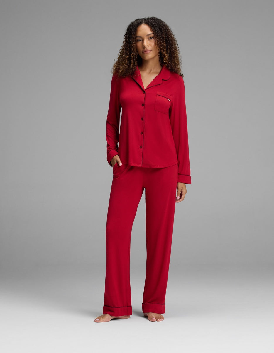 A woman wearing a red Women’s Bamboo Stretch-Knit Long Sleeve Pajama Set, featuring a long-sleeved button-down top with a chest pocket and matching loose-fitting pants, all detailed with black piping.|Color:Cranberry