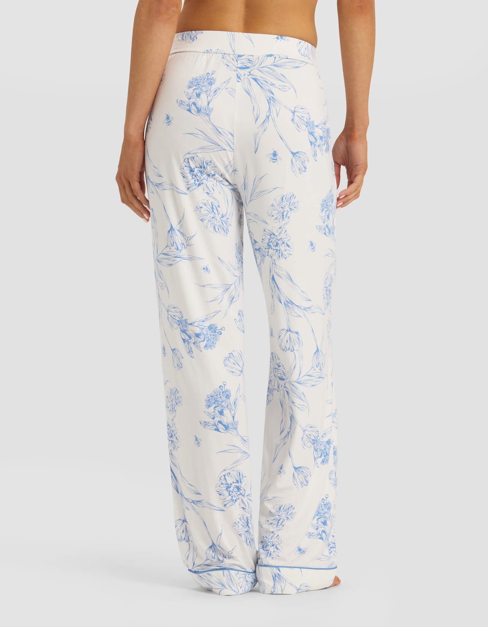 A person is seen from behind wearing Cozy Earth's Women's Bamboo Stretch-Knit Pajama Pant in white with a blue floral print, featuring a loose fit and elastic waistband against a plain white background.