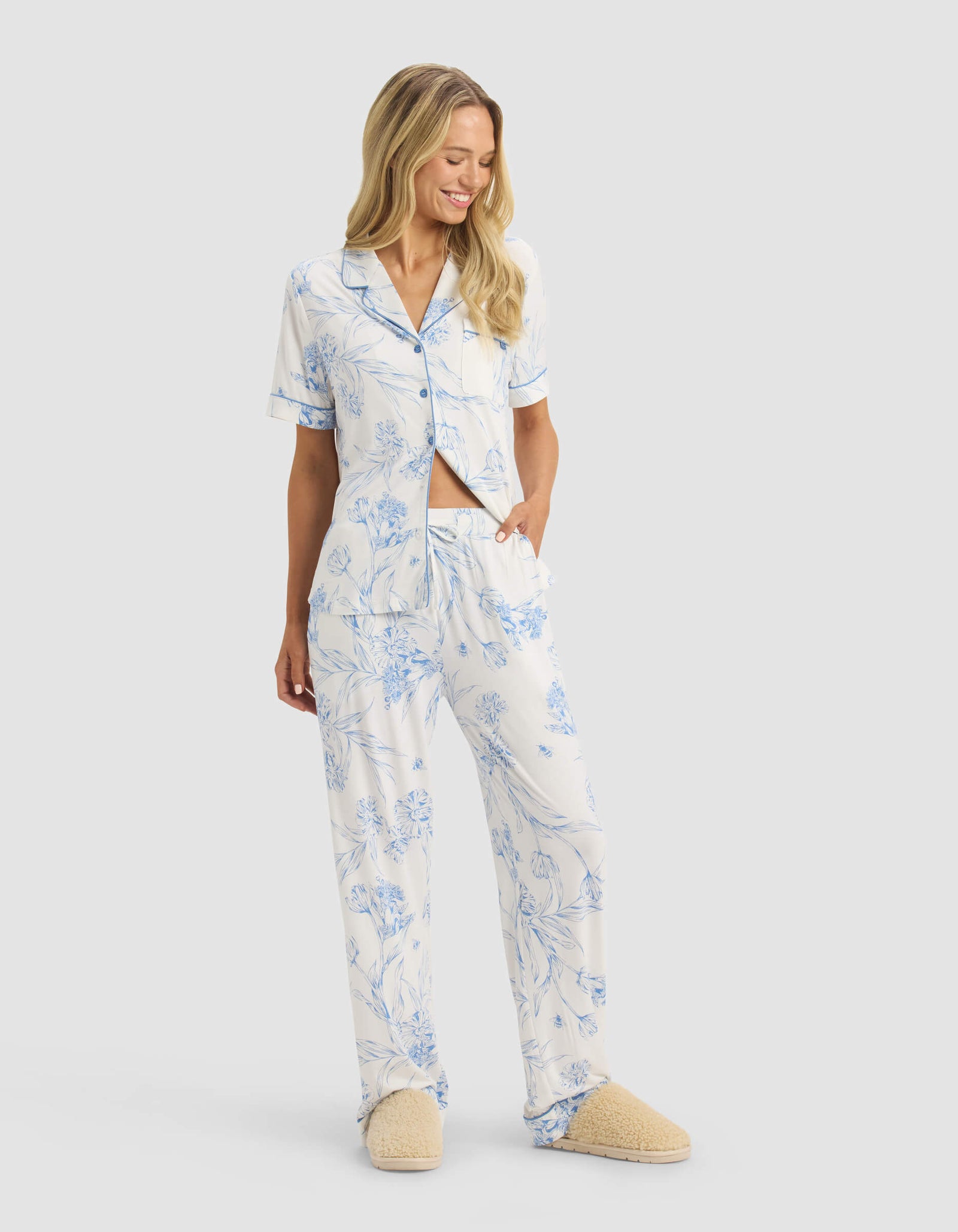 A woman smiles with one hand in her pocket, wearing Cozy Earth's Women's Bamboo Stretch-Knit Pajama Pant and beige slippers, standing against a plain light background.