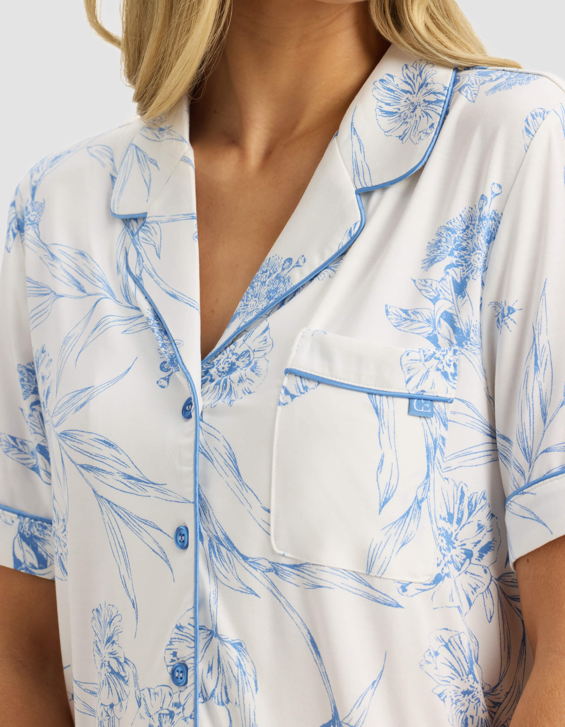 A woman wears the Cozy Earth Women's Bamboo Stretch-Knit Short Sleeve Pajama Top in white, featuring light blue floral patterns, blue buttons, and blue piping on the collar, sleeves, and pocket; only her upper torso is visible.|Color:Garden Toile in Sea Salt