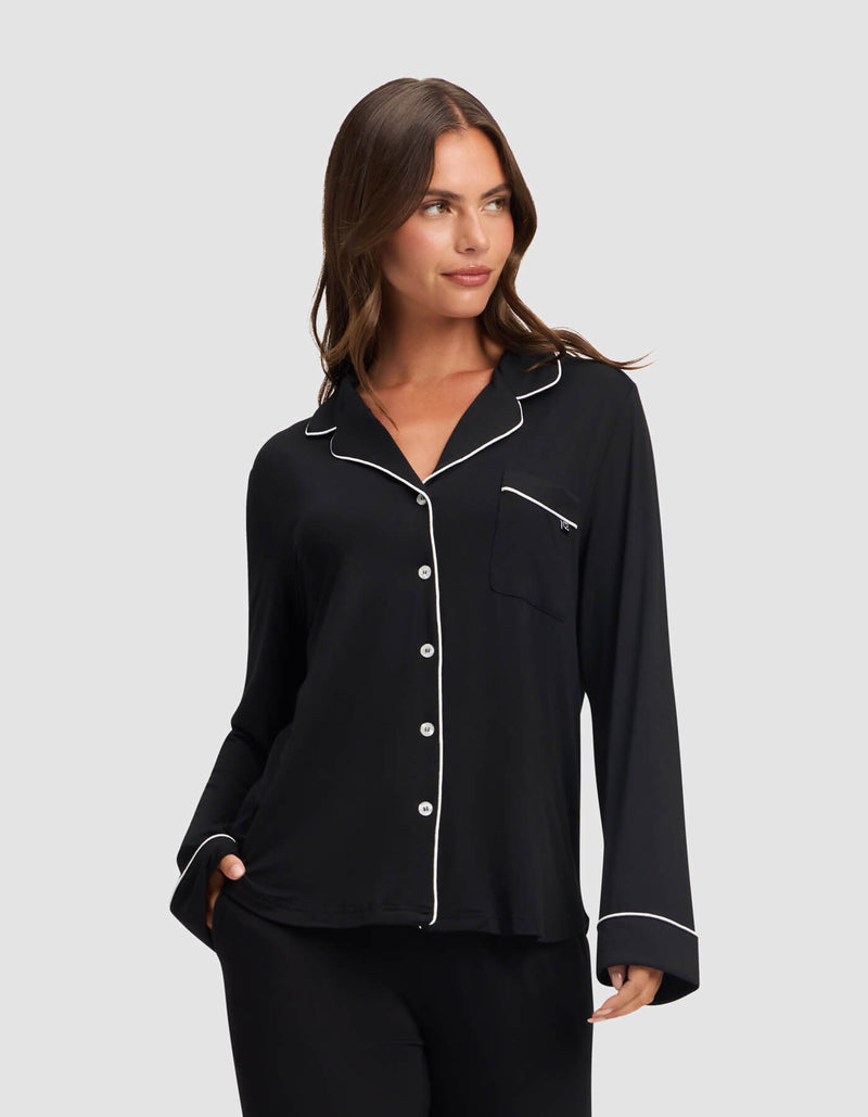 A woman models the Cozy Earth Women's Bamboo Stretch-Knit Long Sleeve Pajama Top in black with white piping on the collar, cuffs, and pocket edges, standing against a light gray background and smiling softly.