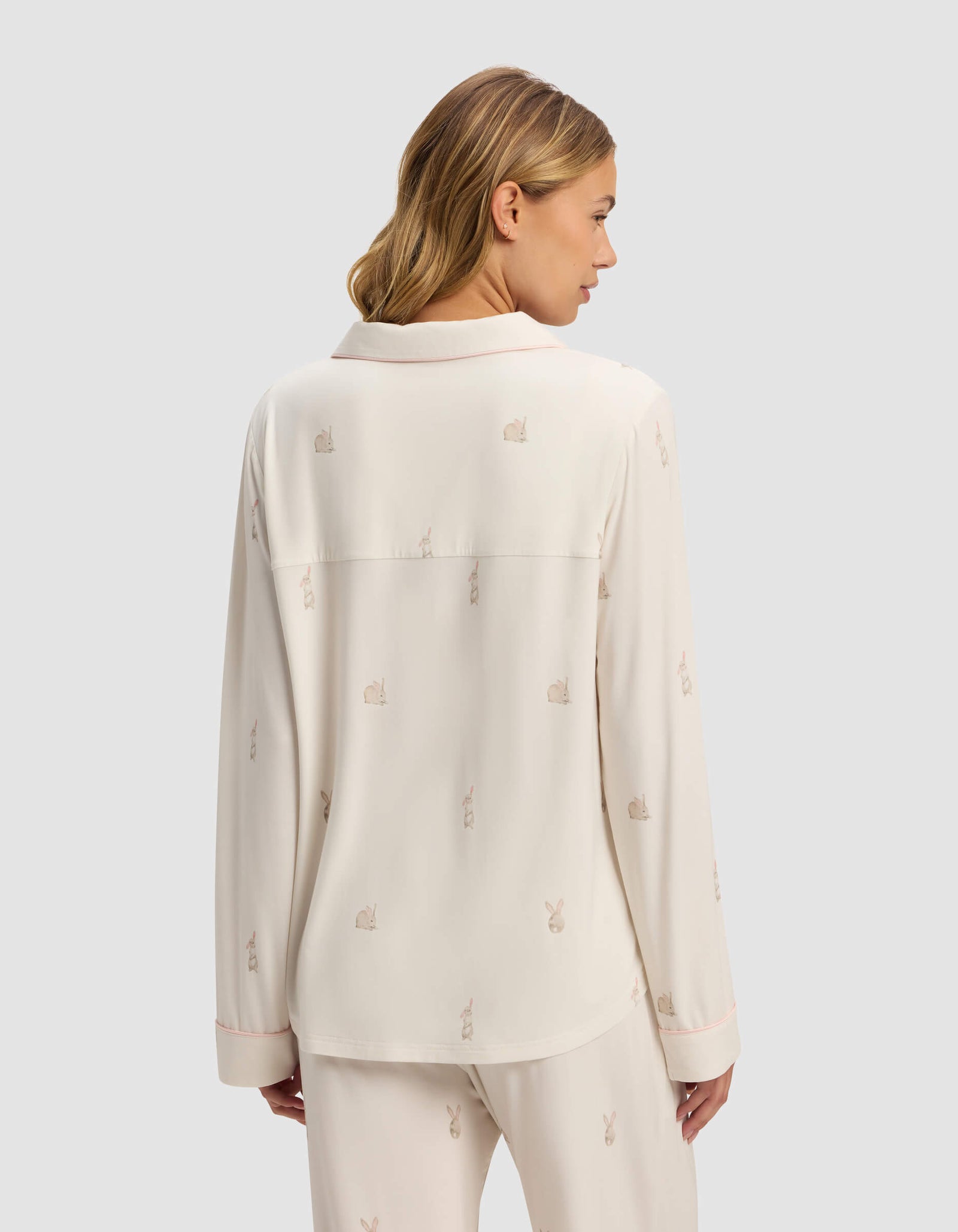 A woman with blonde hair is seen from the back wearing Cozy Earth's Women's Bamboo Stretch-Knit Long Sleeve Pajama Top in cream with a subtle rabbit pattern. The top features long sleeves, a relaxed fit, and is set against a plain light gray background.