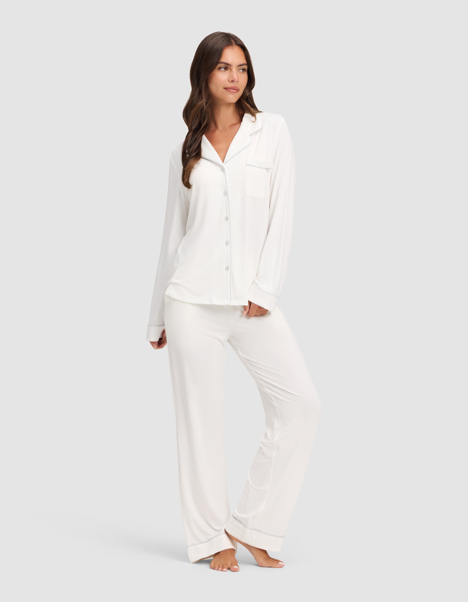 A woman with long brown hair stands barefoot on a light background, wearing the HIDE Women's Bamboo Stretch-Knit Long Sleeve Pajama Top with buttons and a chest pocket, and poses with one hand in her pocket.
