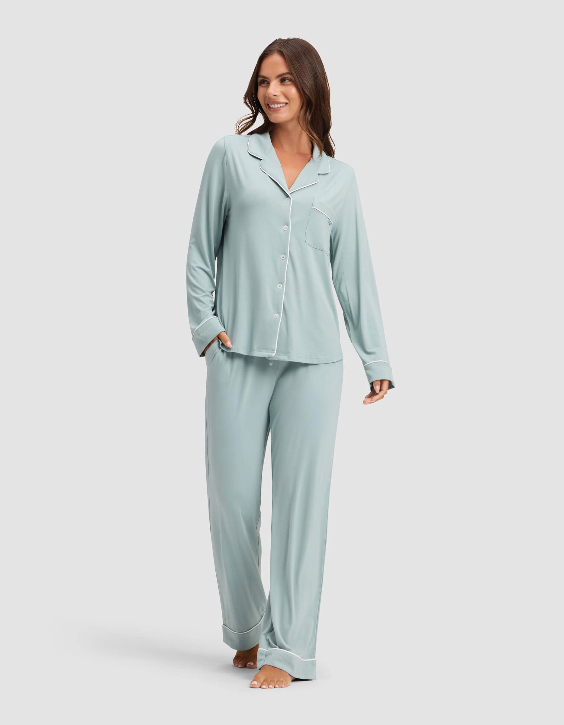 A woman smiles while wearing Cozy Earth Women's Bamboo Stretch-Knit Pajama Pant in light blue with a matching long-sleeve button-up top, barefoot against a plain light background. |Color:Brook