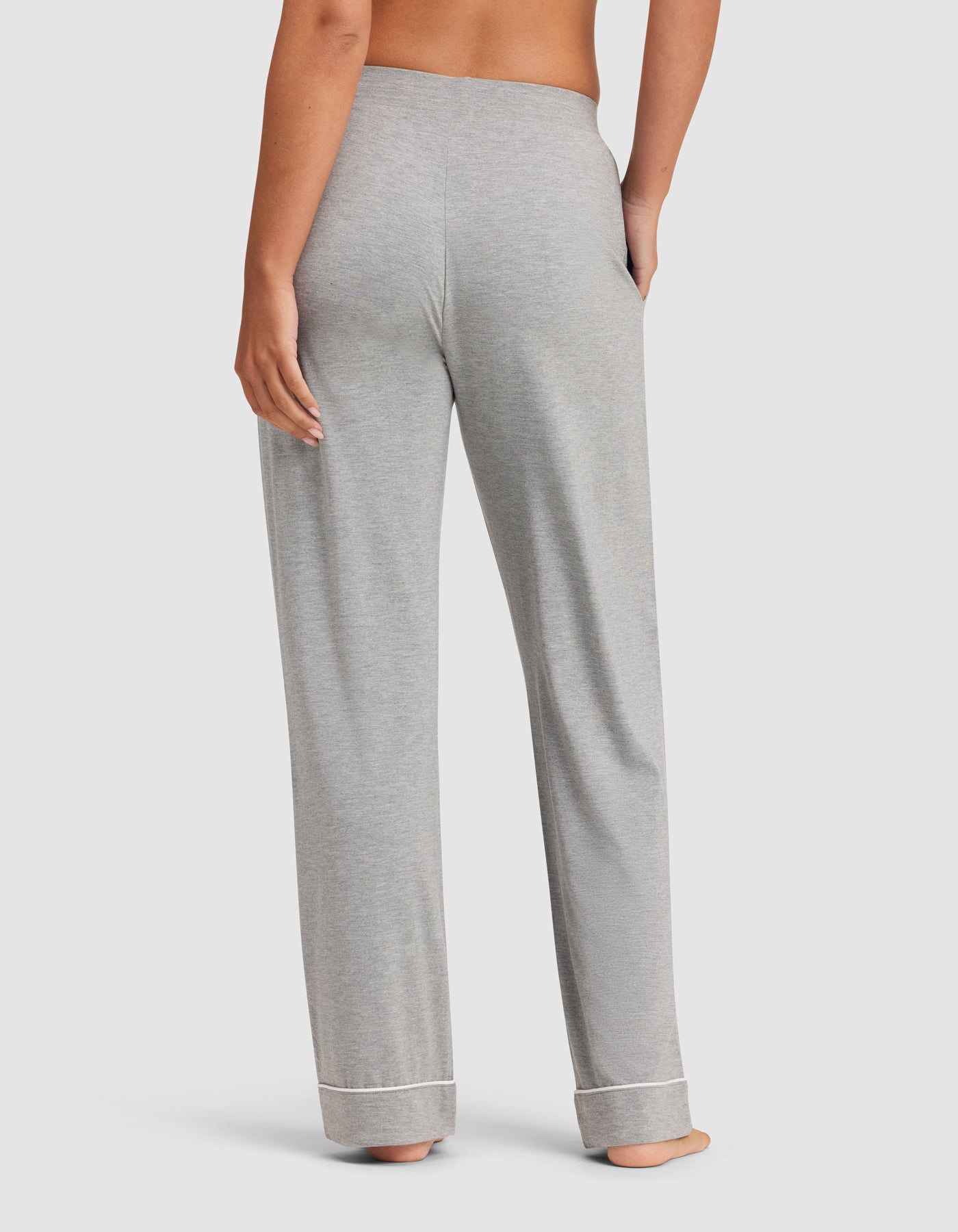 A person stands barefoot with their back to the camera, wearing HIDE Women's Bamboo Stretch-Knit Pajama Pants in light gray with a loose fit, white trim at the cuffs, and their right hand in a pocket.
